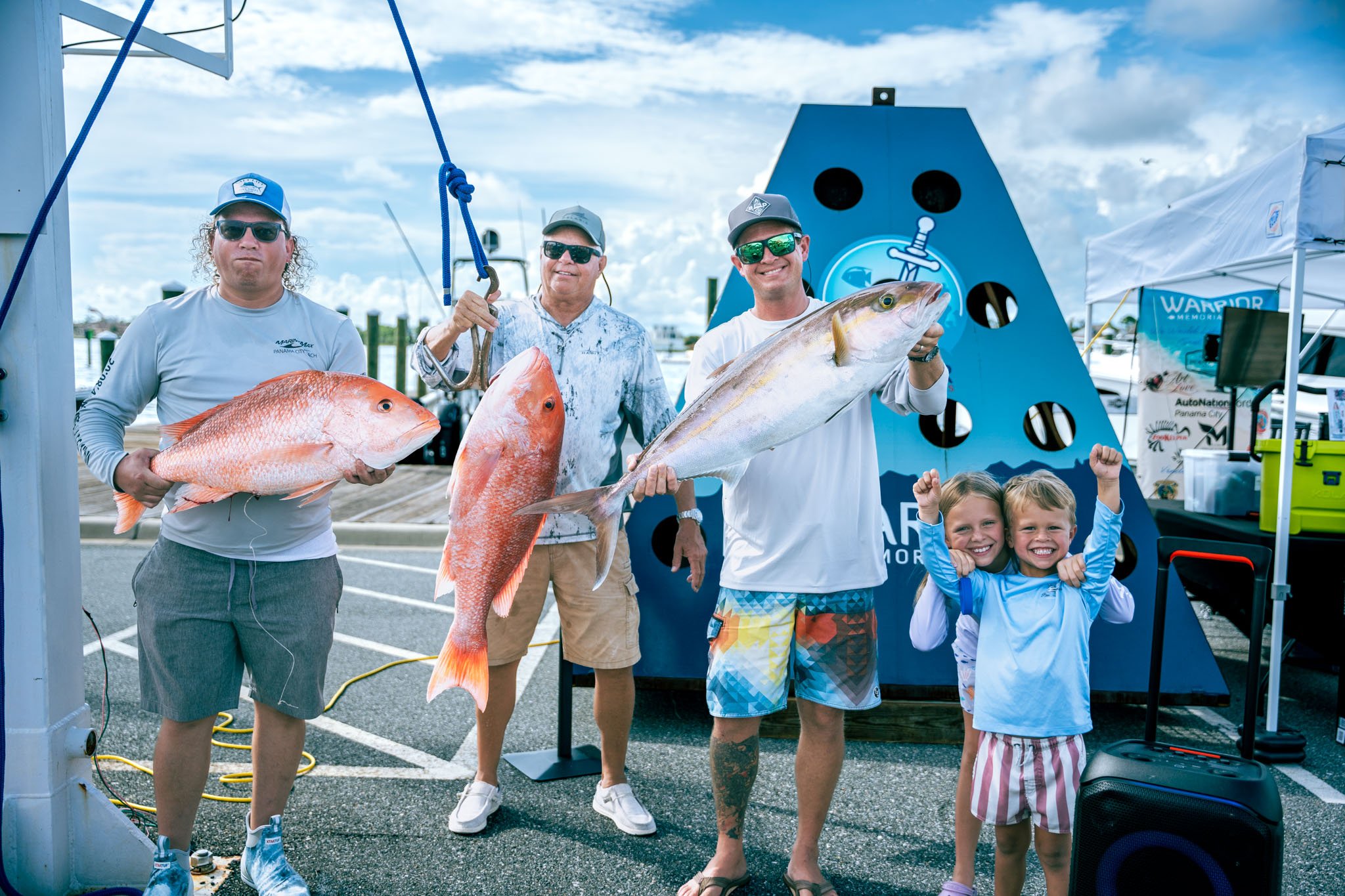 Three men and two children at a seafood dock, holding large fish they caught, with a blue boat and banner in the background at Point South Marina - Bay Point (Panama City Beach).
