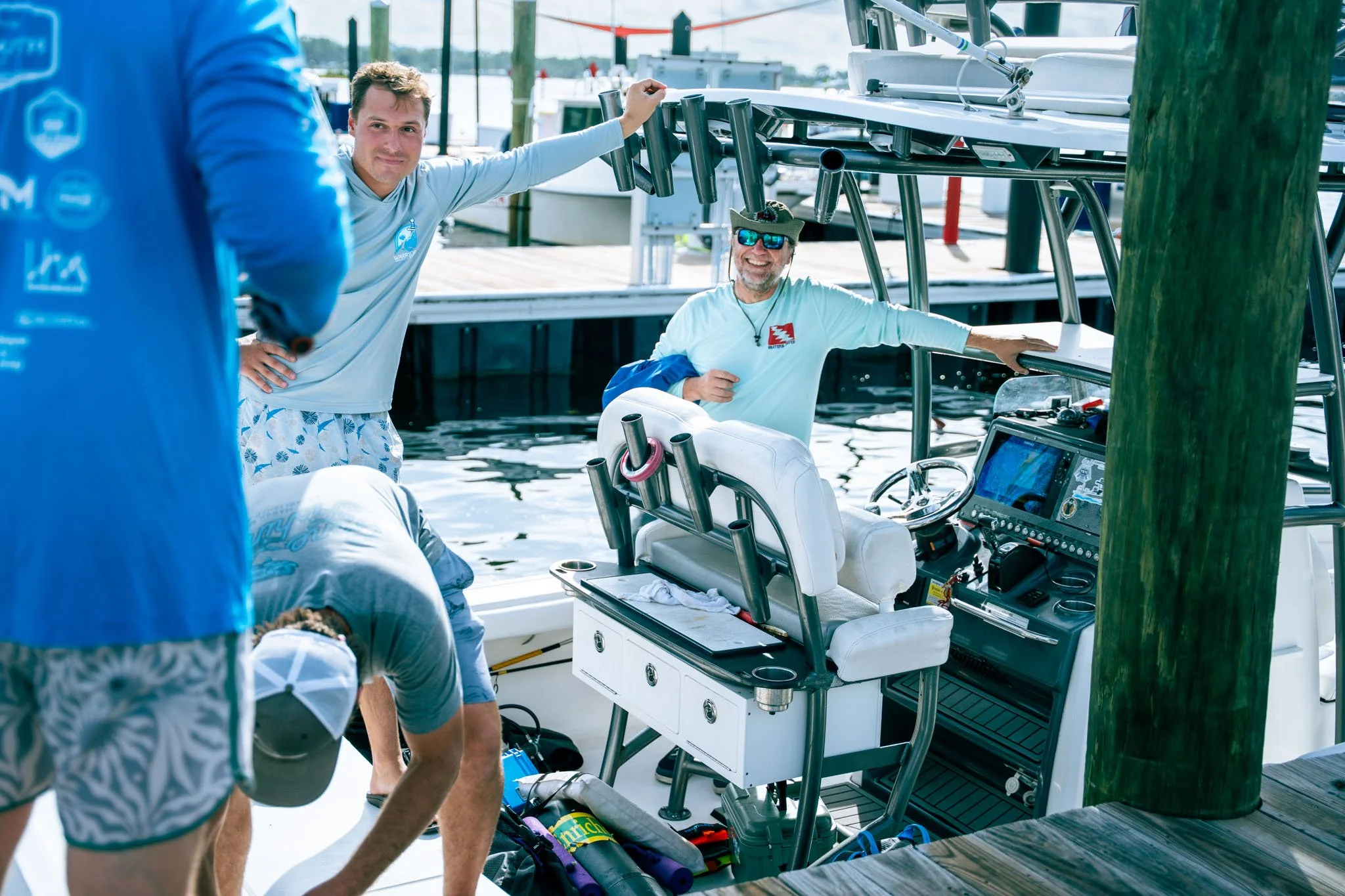 Two men on a boat preparing fishing equipment, with one man sitting on an elevated seat and smiling, and the other man partly visible in the foreground.