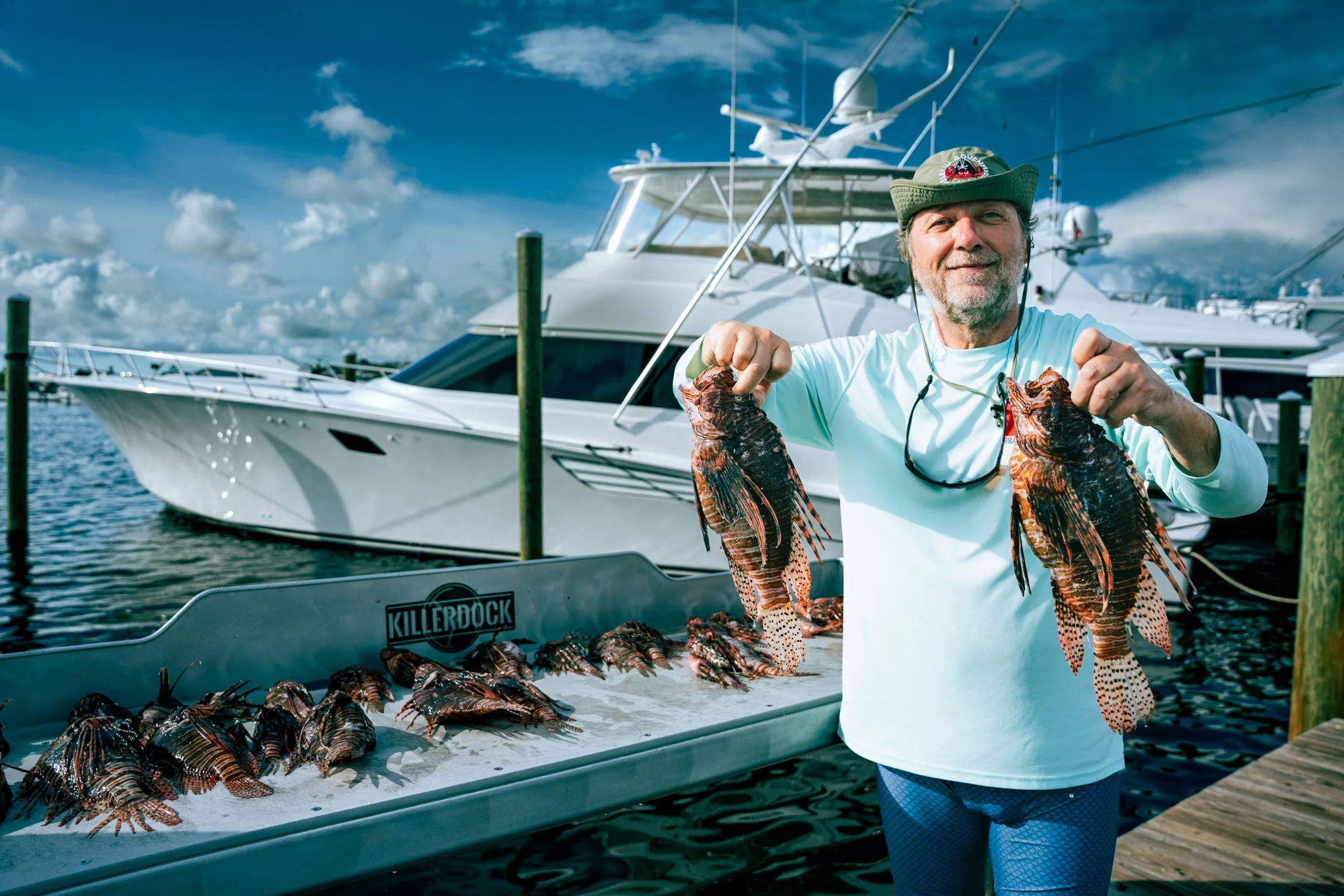 A man holding two large lionfish in front of a boat docked at Point South Marina - Bay Point (Panama City Beach), with more lionfish on the waterfront cleaning station surface, and yachts visible in the background under a partly cloudy sky.
