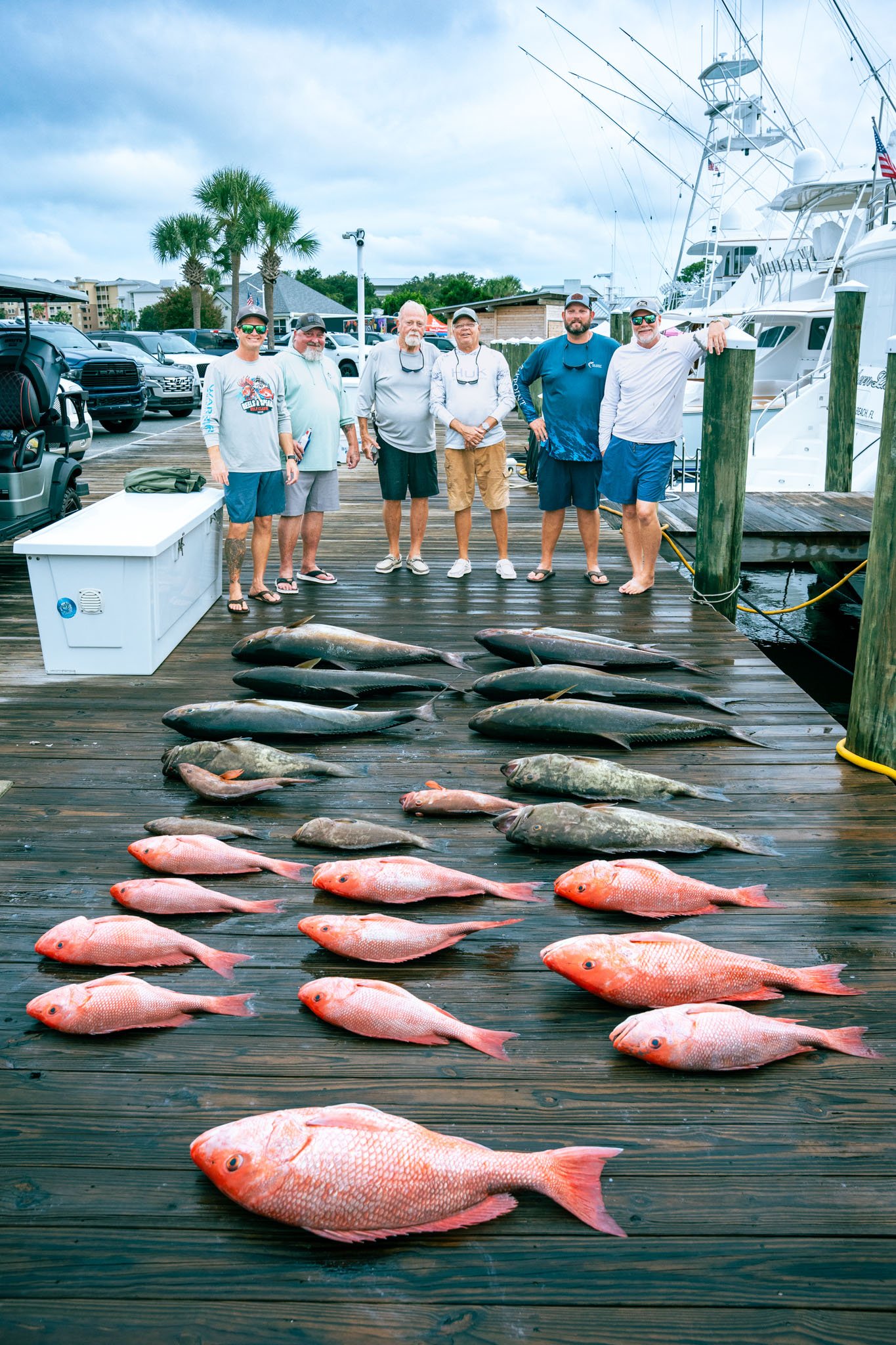 A group of six men standing on a dock with boats in the background, several fish laid out in front of them, including red and gray-colored fish.