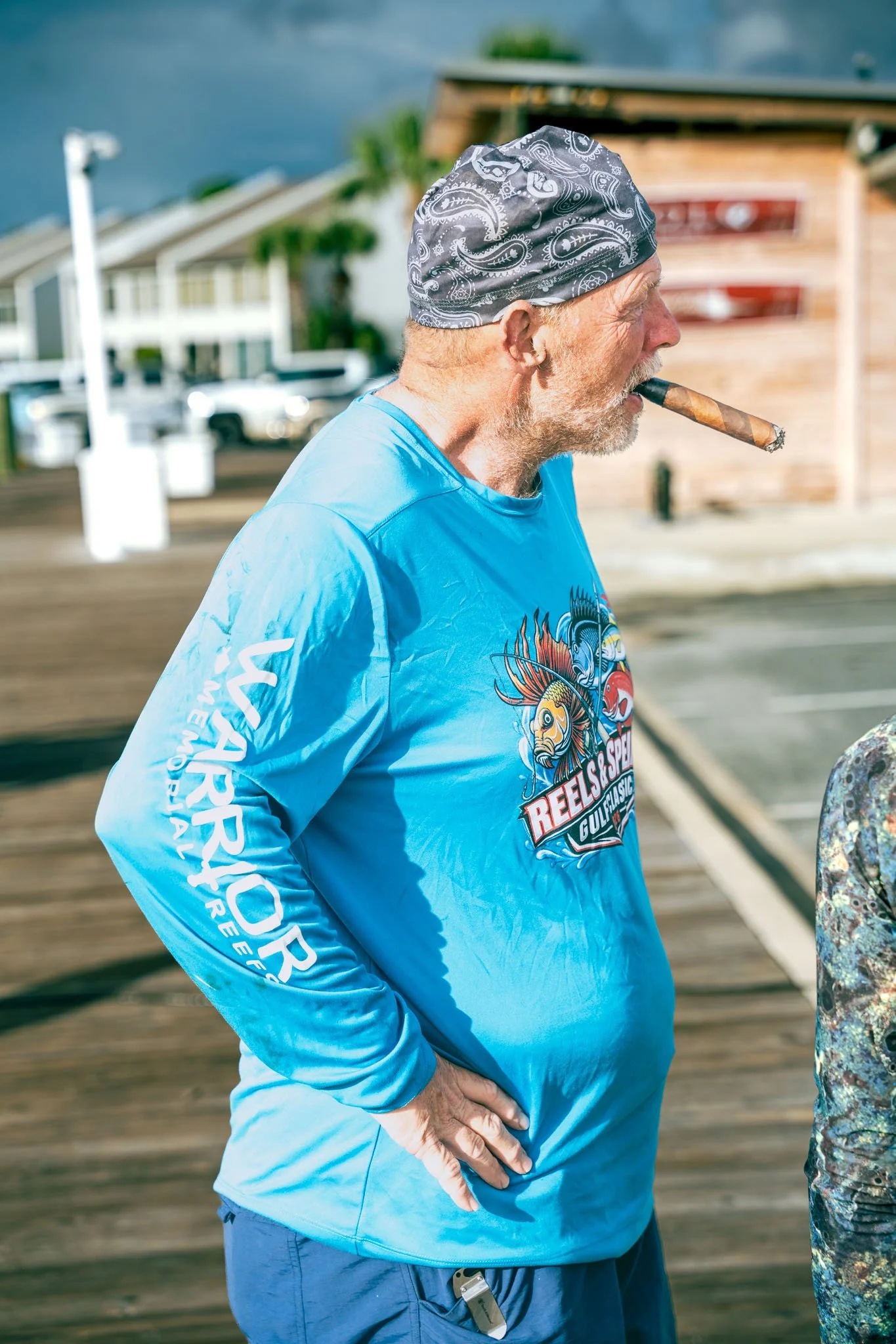 An older man with a gray beard, wearing a gray bandana and a light blue long-sleeve shirt with a graphic design, is smoking a cigar on a wooden pier near water. Another person partly visible on the right is wearing camouflage clothing.