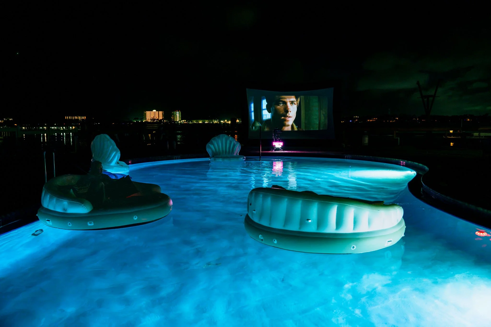 Nighttime scene of Point South Marina - Bay Point (PCB, FL) poolside movie screening, with floating inflatable loungers illuminated by blue light, a large outdoor screen showing a man's face, and city buildings with lights in the background.