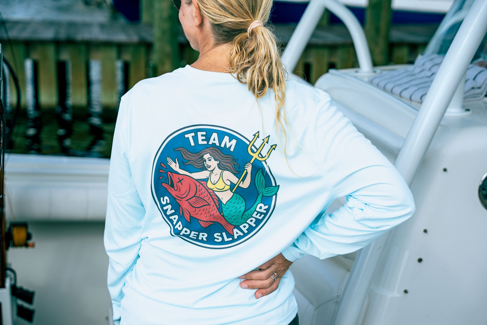 Back of a person with blonde hair tied in a ponytail, wearing a white jacket with a colorful emblem that includes a mermaid holding a trident and the words 'Team Mermaid Snapper Slapper' on it. Person is standing outdoors near a white outdoor bench.