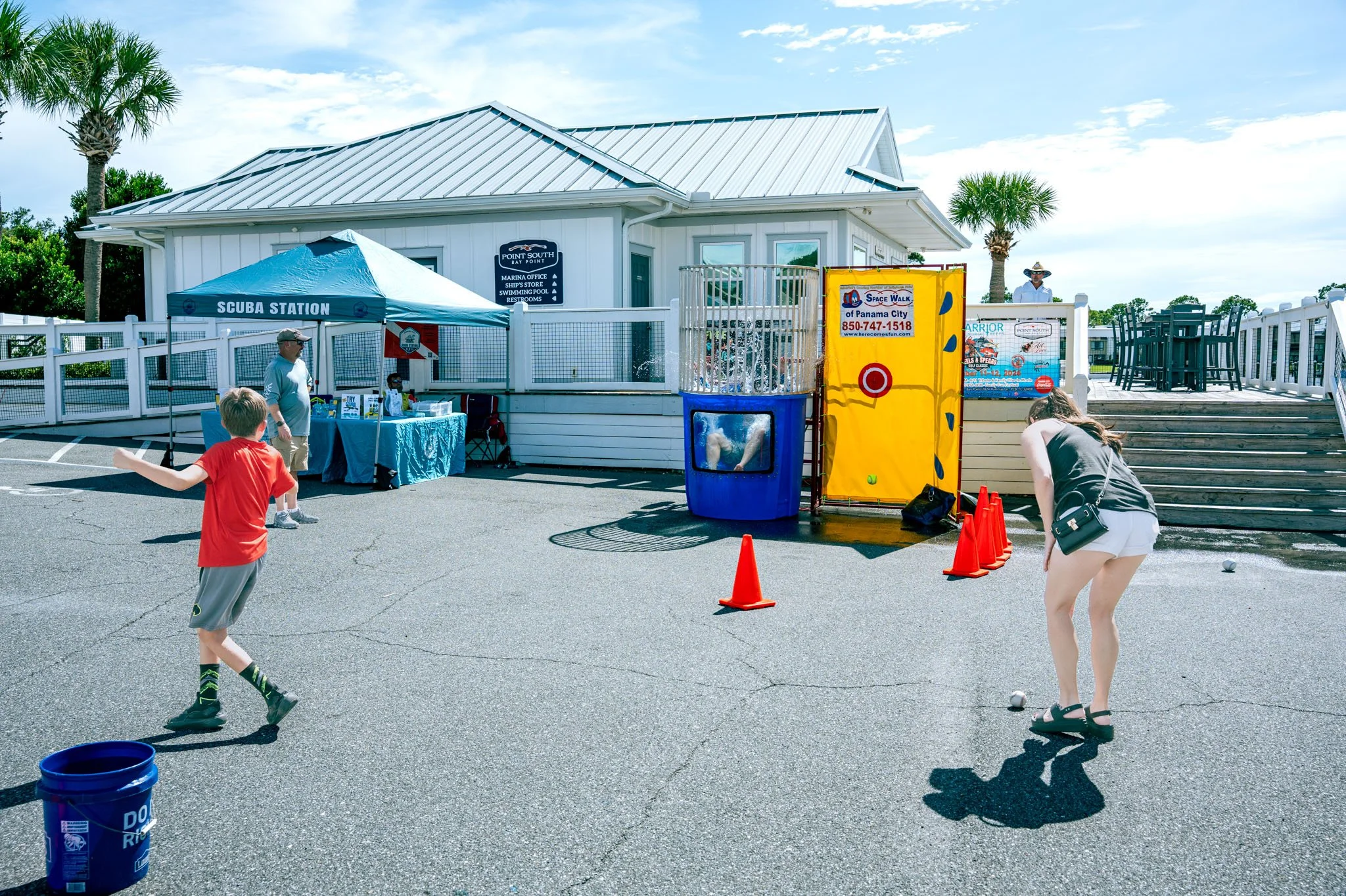 People playing a game of bocce ball or similar outdoor game on a paved area near a white building with a sign, a tent labeled 'SCUBA STATION,' and orange cones shaping the area for the game, against a backdrop of palm trees and a partly cloudy sky.
