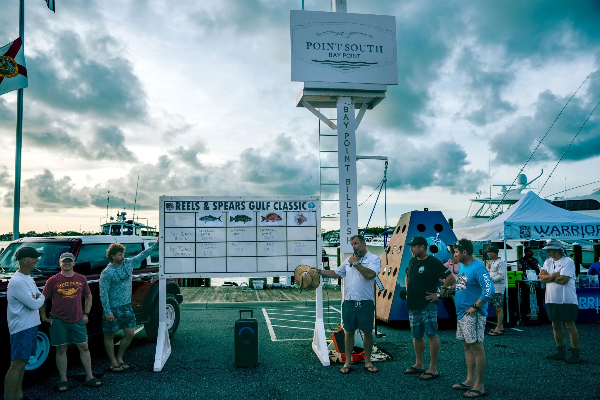 People gathered at the Gulf Classic fishing event registration area, with a sign listing winners for reel and spear categories, boats in the background, and cloudy sky overhead.