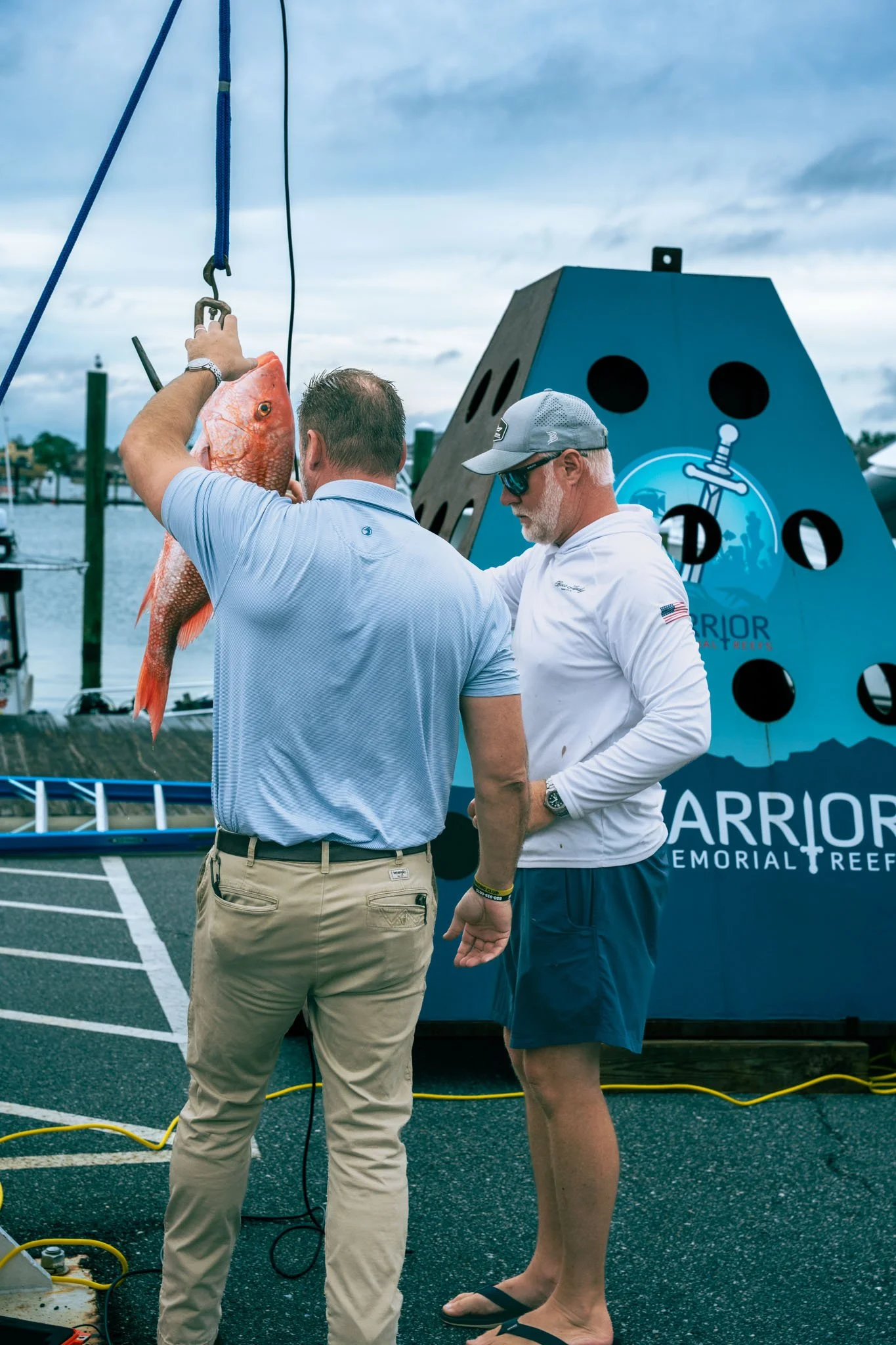 Two men are involved in a scuba diving event, one holding a red fish, standing on a parking lot with a memorial reef advertisement in the background and cloudy sky overhead.