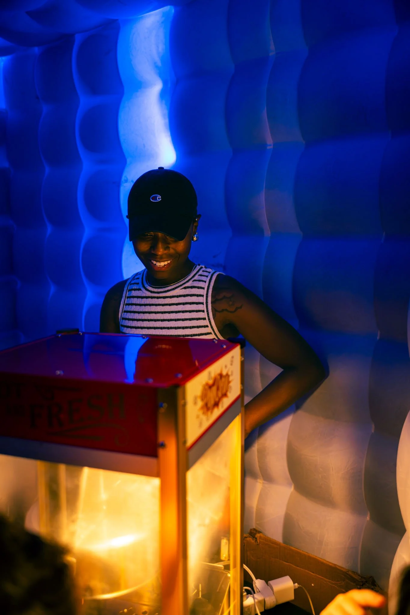 A smiling Black woman wearing a black face cap with a Champion logo, striped sleeveless top, and earrings, standing behind a popcorn machine with a red and yellow sign on it, against a textured blue wall background.
