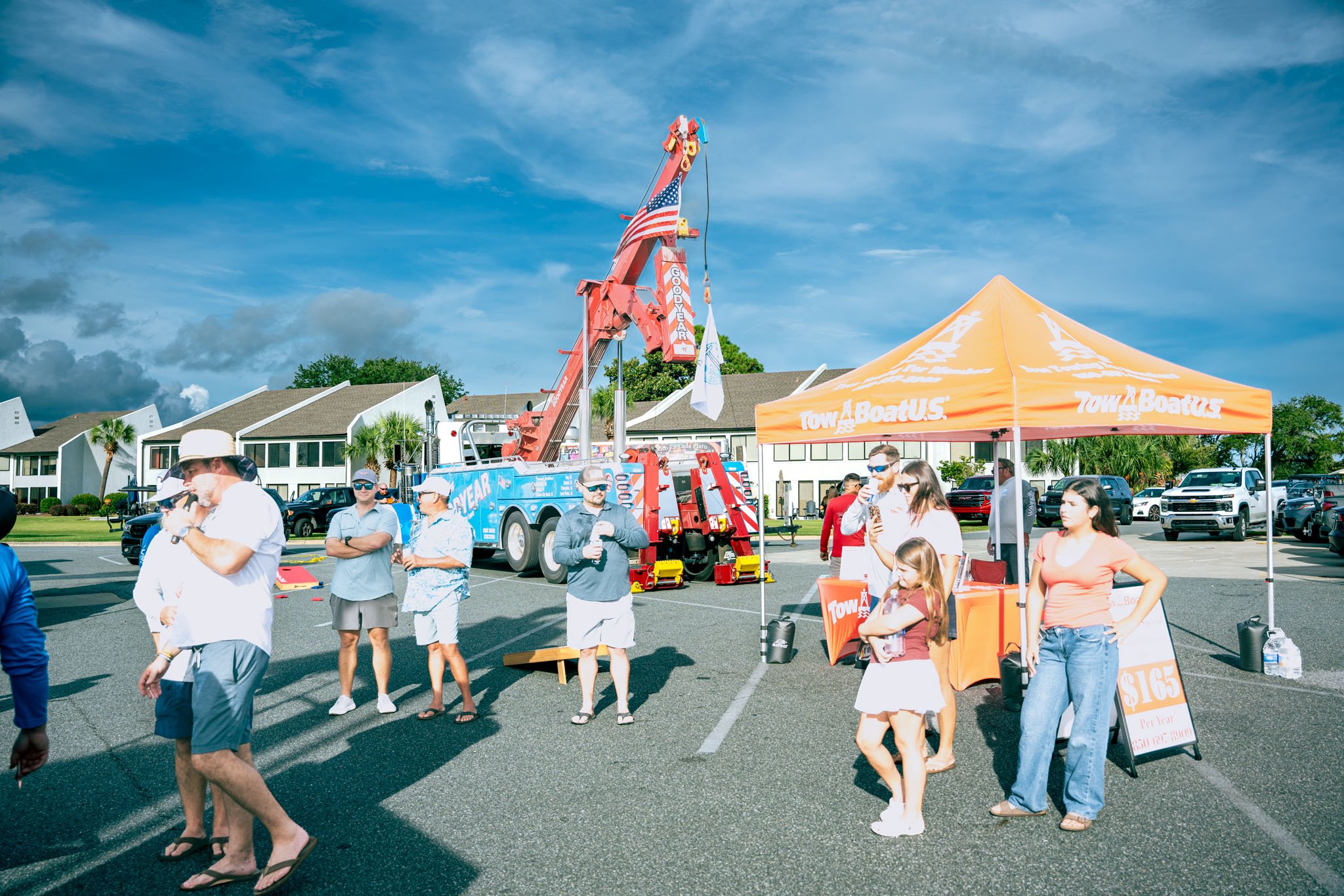 People gathered around a large tow truck with a crane, at an outdoor event. There is a bright orange tent with the Tow A Boat US logo, and a banner displaying a special offer. The background shows residential buildings, palm trees, and a partly cloud