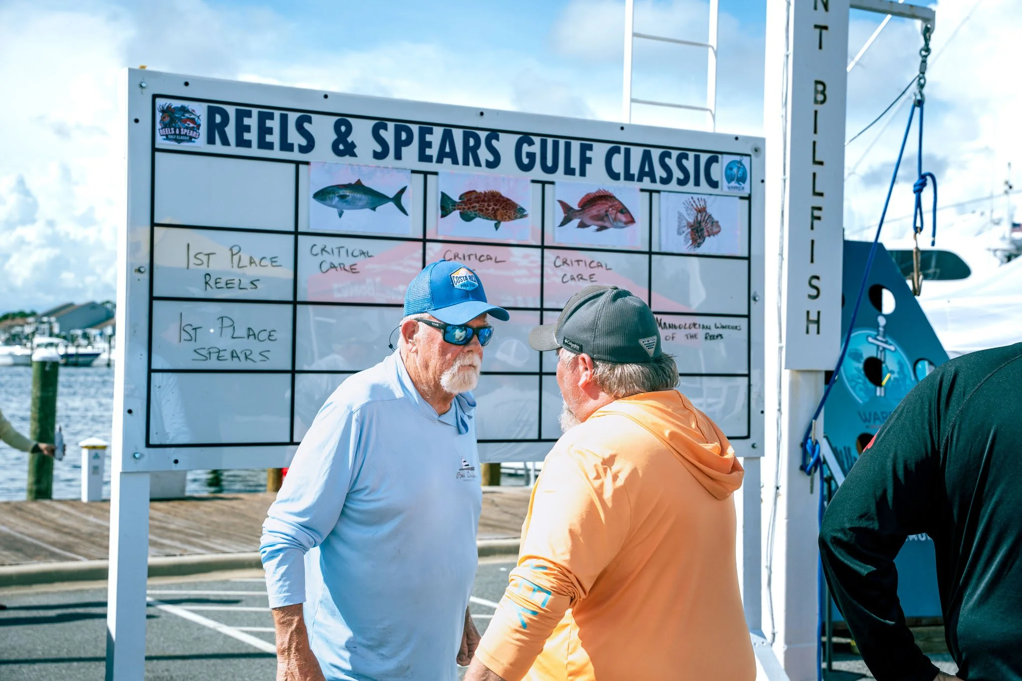 People standing near a scoreboard at a fishing competition, with charts for species and placements, noting critical care and winners for reels and spears at the Reels & Spears Gulf Classic event.