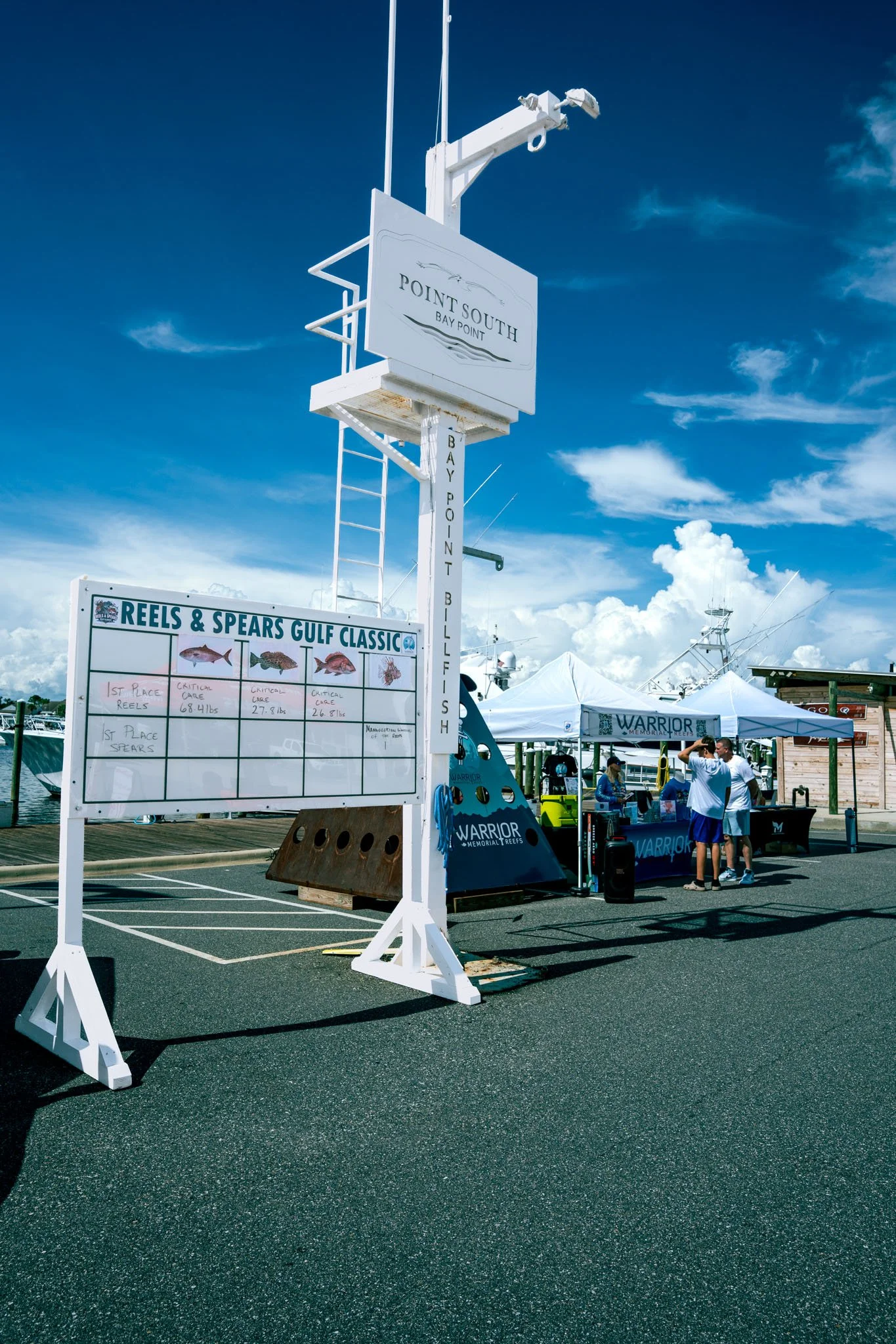 Point South Marina - Bay Point (Panama City Beach) with a sign for Point South Bay Point, and a white tent setup at a fishing event called Reels & Spears Gulf Classic, with boats and people in the background under a partly cloudy sky.