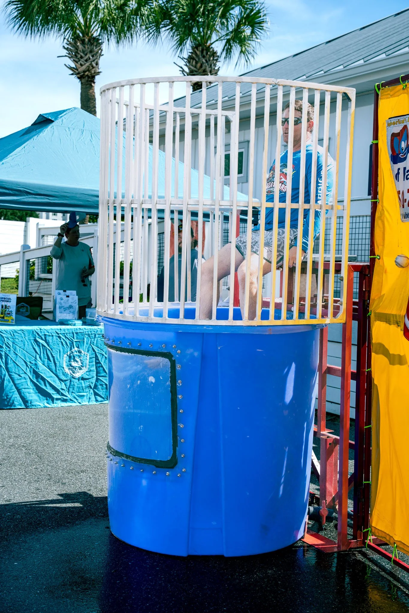 A person in a blue and white swimming shirt and shorts sitting inside a tall, blue cylindrical water tank with white vertical bars, during daytime at an outdoor event. Palm trees, a white house, and a yellow banner with a cartoon character are visibl