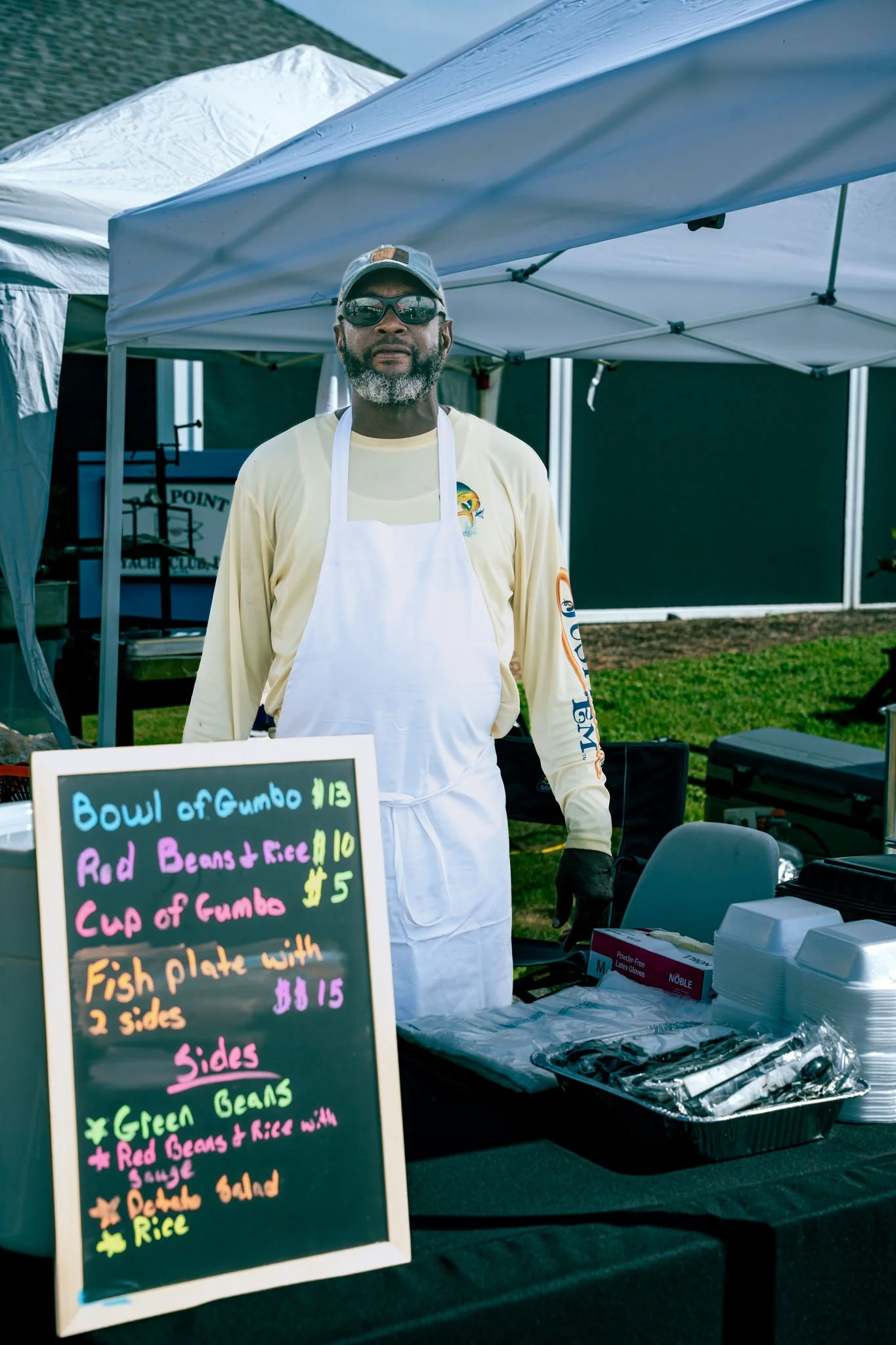 A man with sunglasses and a beard stands behind a food stall at an outdoor event. The stall features a colorful menu board listing dishes including bowls of gumbo, red beans and rice, and fish plate with sides. The man is wearing a yellow long-sleeve