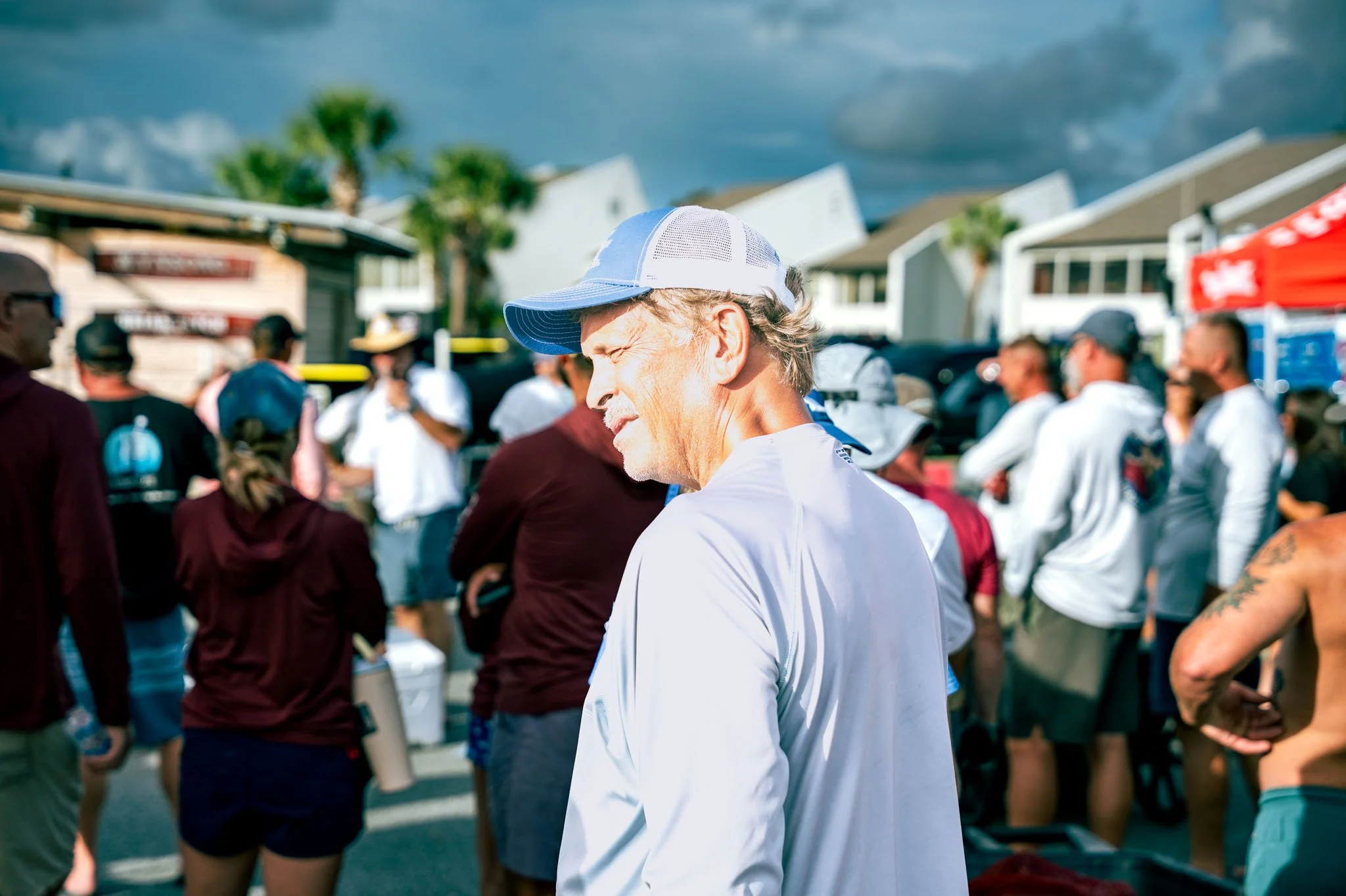 A man with a gray beard and mustache wearing a white shirt and a blue and white baseball cap, with his eyes closed, standing among a crowd of people outdoors on a cloudy day.