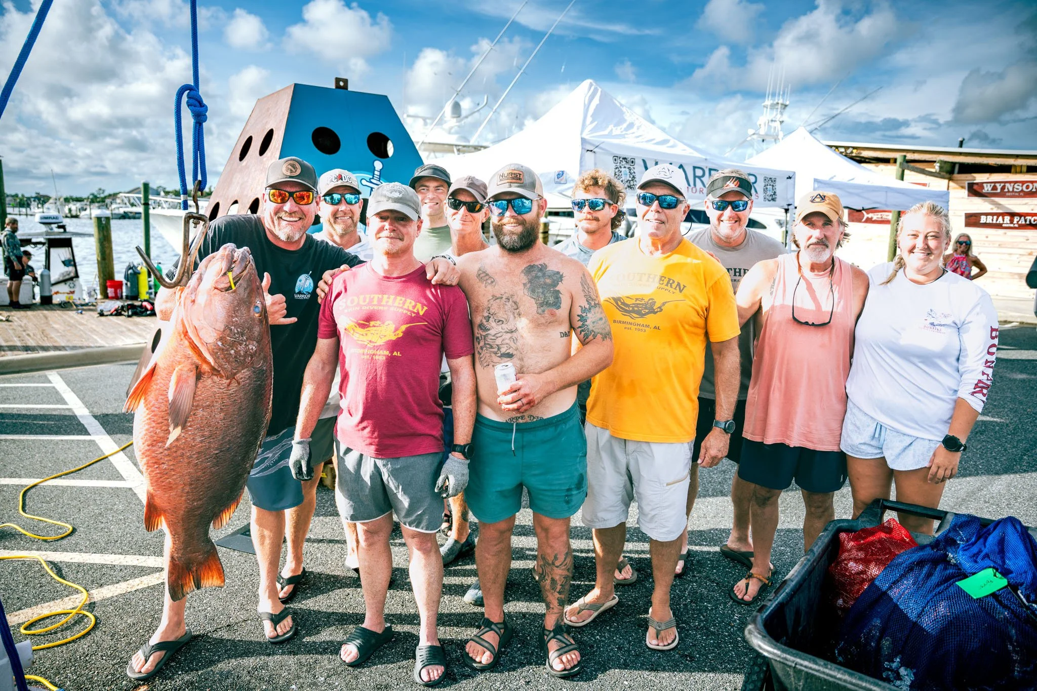 Group of people at a marina holding a large cubera snapper, with tents, boats, and a blue sky in the background.