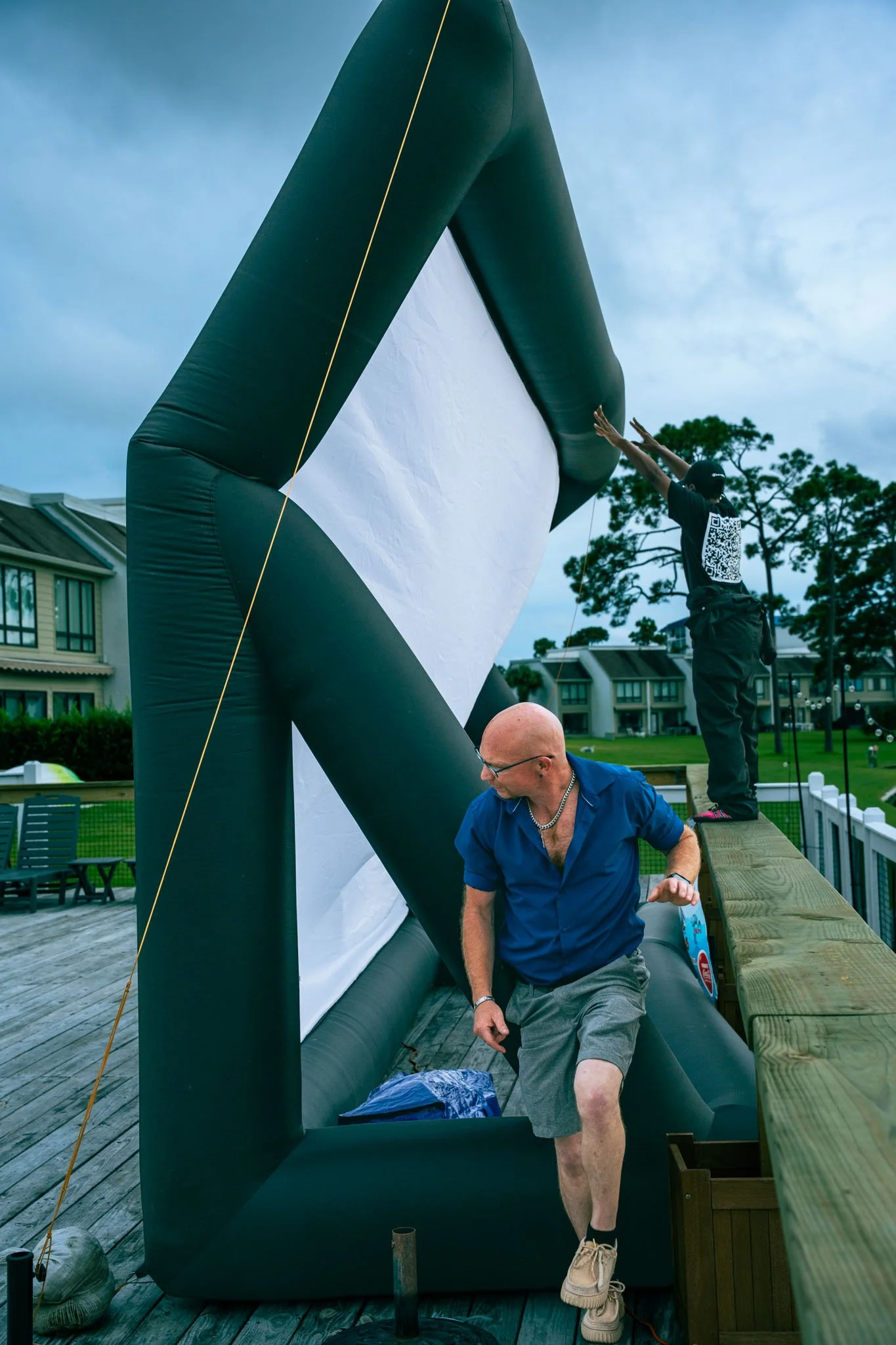 Two men are assembling or working on a large, inflatable black and white structure outdoors on a wooden deck, with houses and trees in the background.