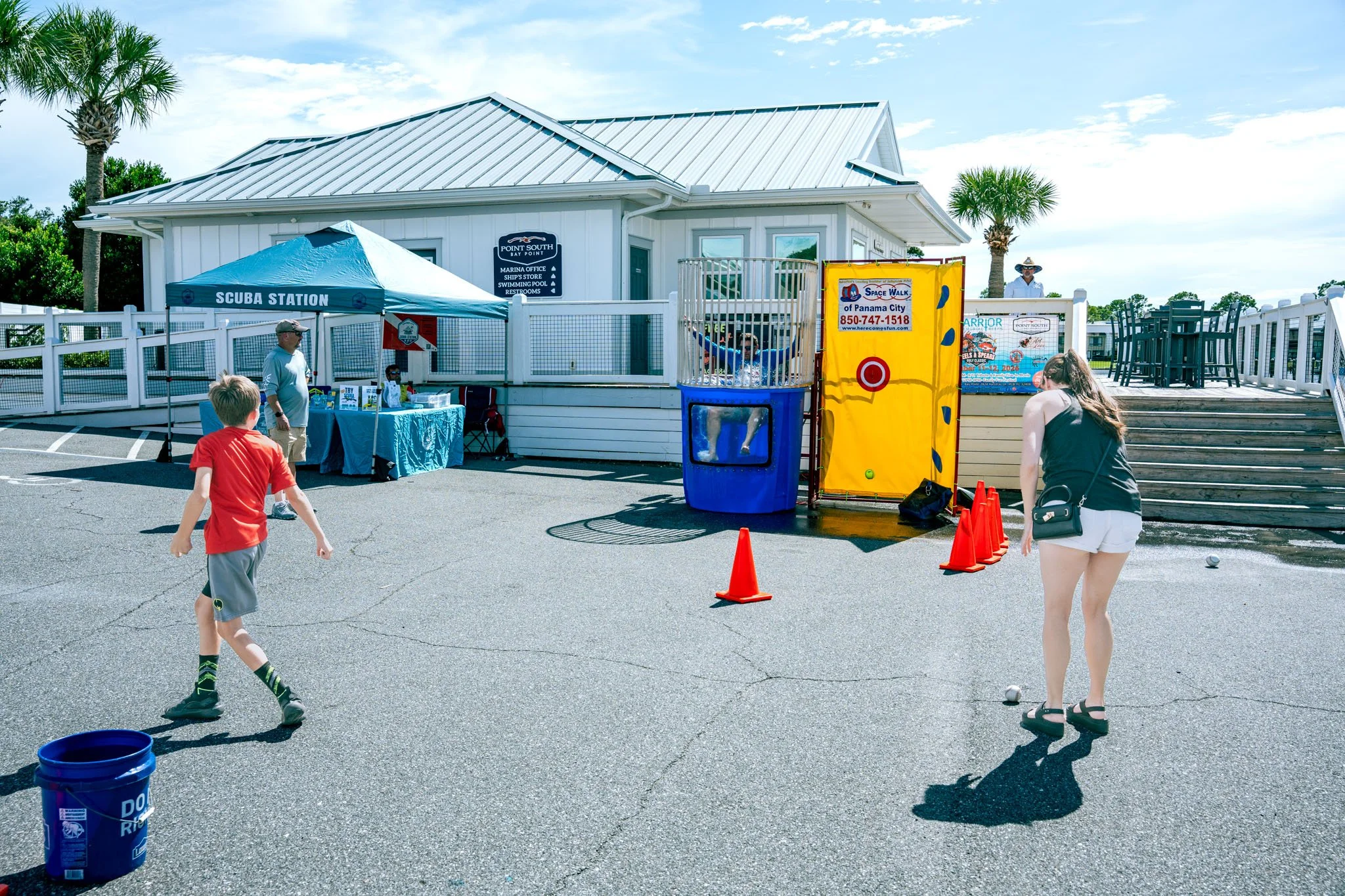 People playing a game of bocce ball outside near a white building with a blue sign that reads 'Point South Bay Point'. Children and adults are present, with some children running and playing, and one person observing the game.