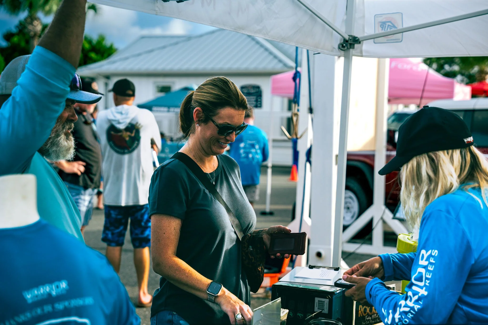 People interacting at the Warrior Memorial Reefs Reels and Spears Fishing Tournament event at Point South Marina with tents and vehicles in the background, one woman smiling at a table with a camera, others are wearing sunglasses and casual clothing.