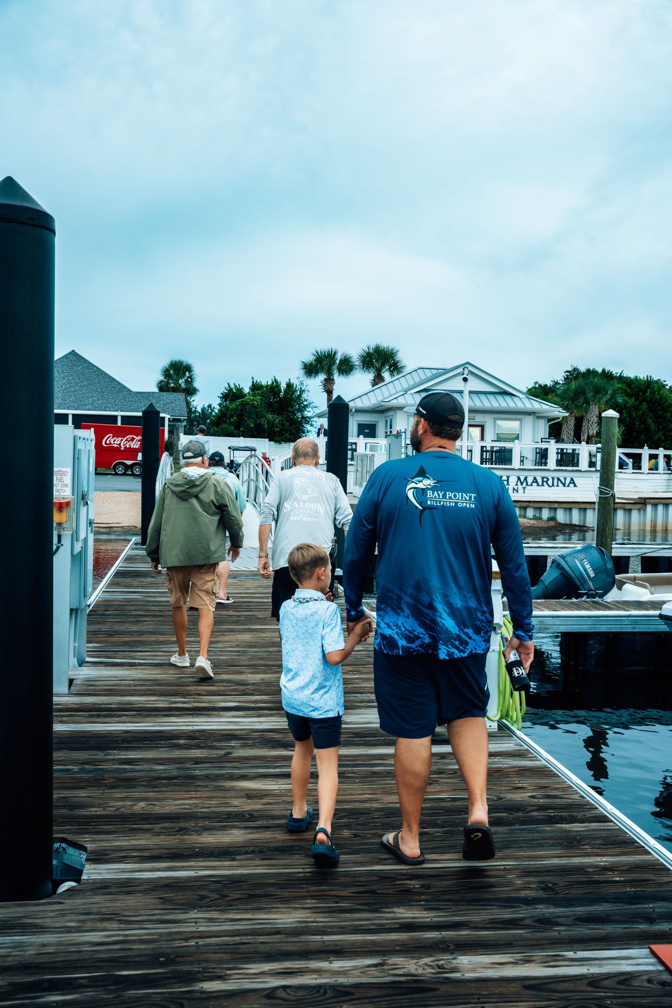 People walking along a wooden dock at Point South Marina - Bay Point (Panama City Beach) towards the marina with boats and buildings, some palm trees, overcast sky.