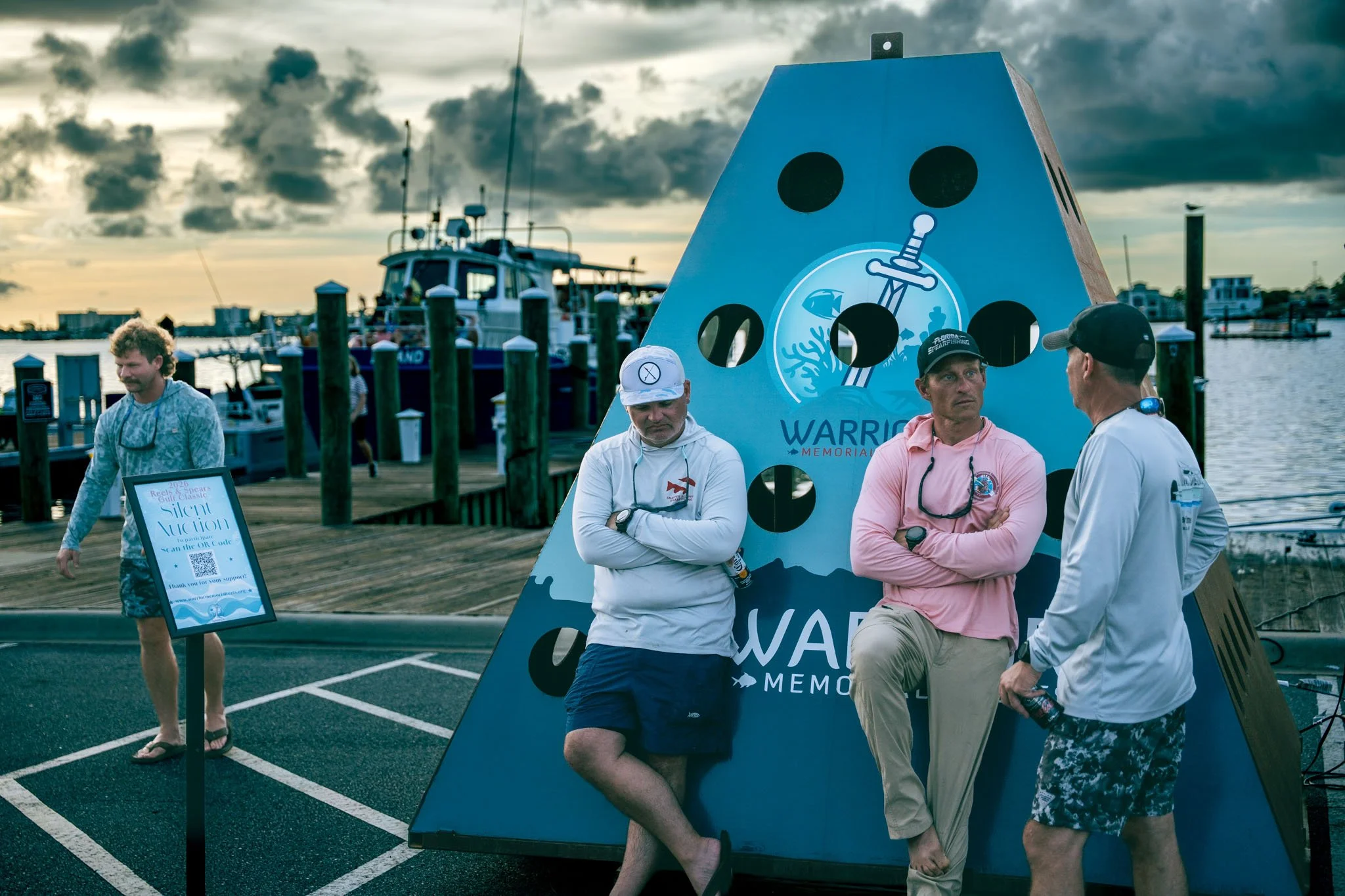 A scene at a marina or dock with people gathered around a memorial display. The display features a large blue board with black dots, a dolphin and coral logo, and text that reads "Warrior Memorial." Several men are conversing near the display, some w