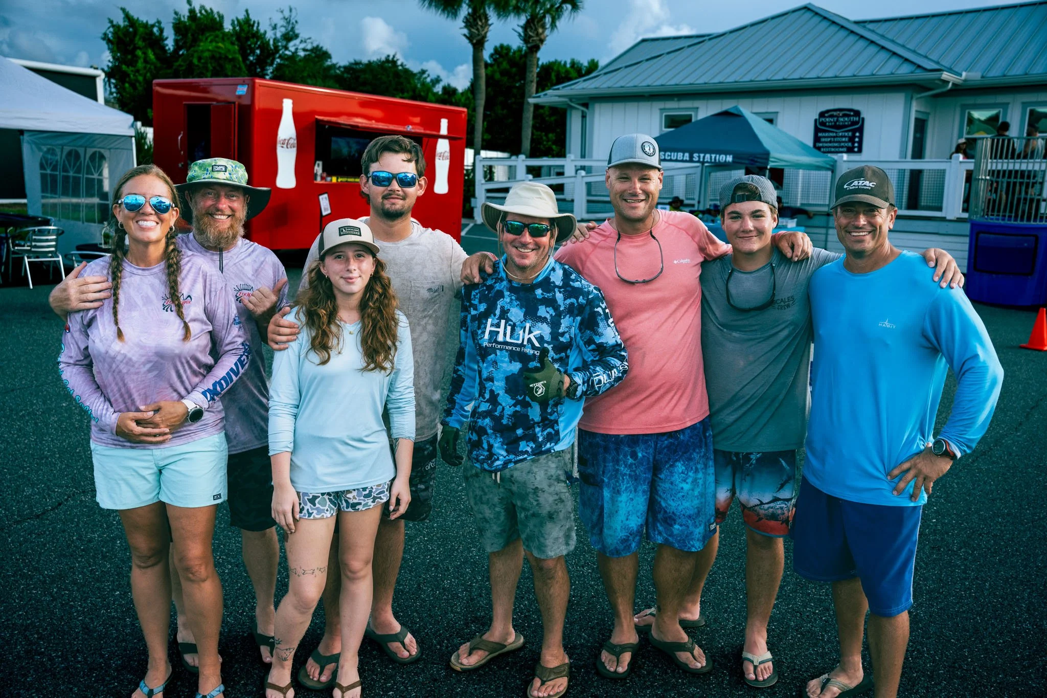 A group of nine people standing outdoors in casual summer clothing, smiling, with palm trees, a building with a sign that reads 'Scuba Station,' and a red Coca-Cola vending trailer in the background.