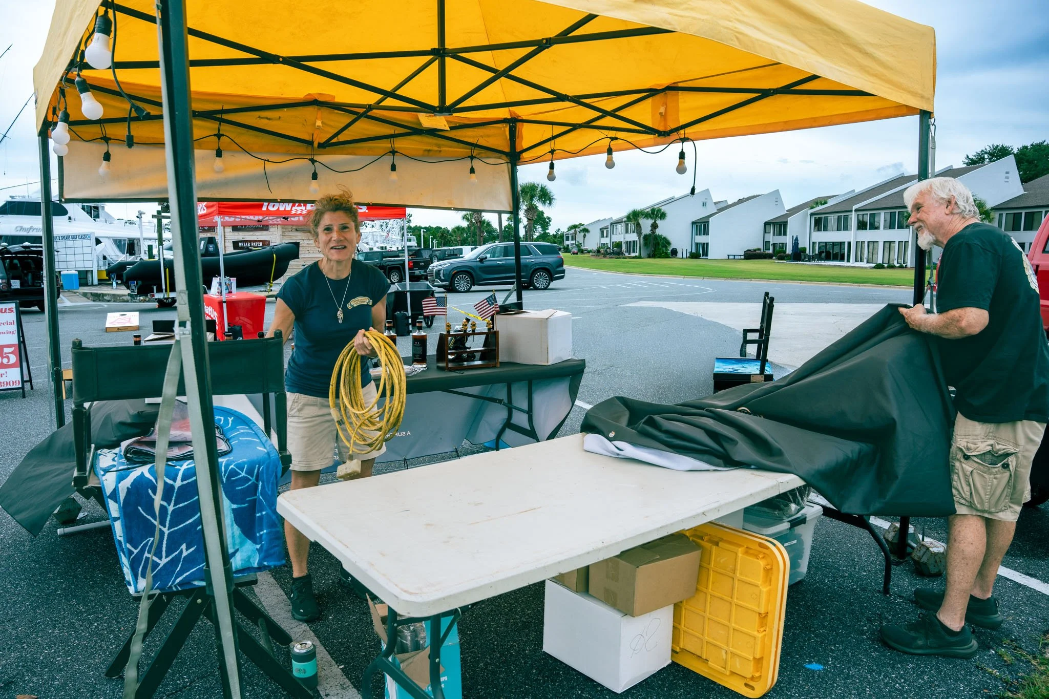 A woman is standing behind a table under a yellow canopy at an outdoor market, holding a yellow hose. A man is on the right side, partially visible, handling a large black bag. The background shows parked cars, buildings, palm trees, and a cloudy sky