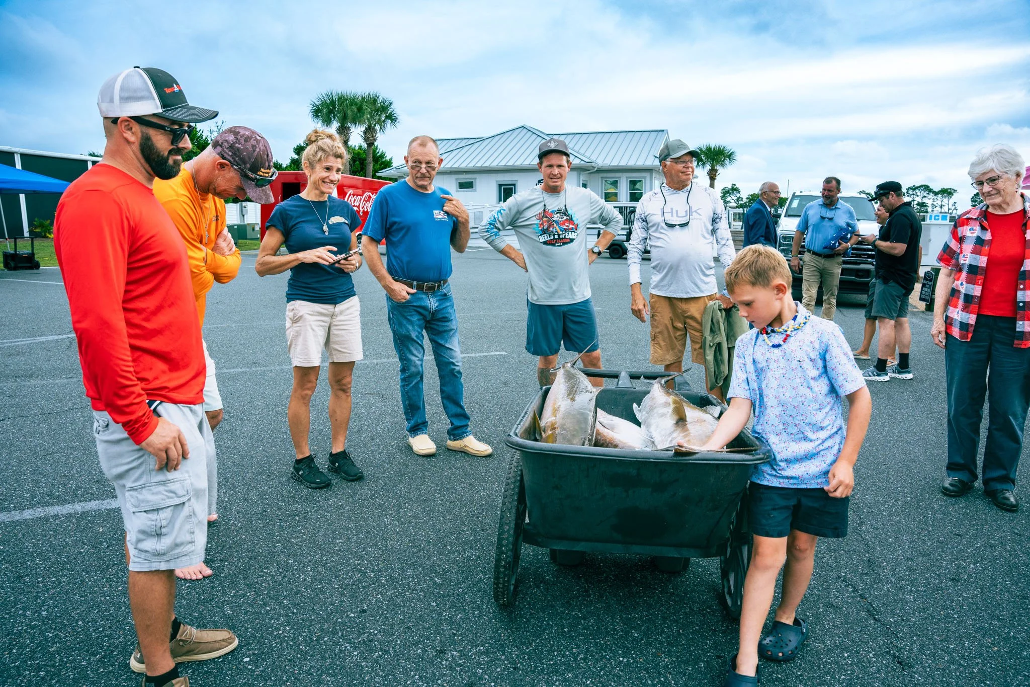 A group of people gathered outdoors around a container with large fish, possibly after a fishing event, on a paved surface with a building, palm trees, and vehicles in the background.
