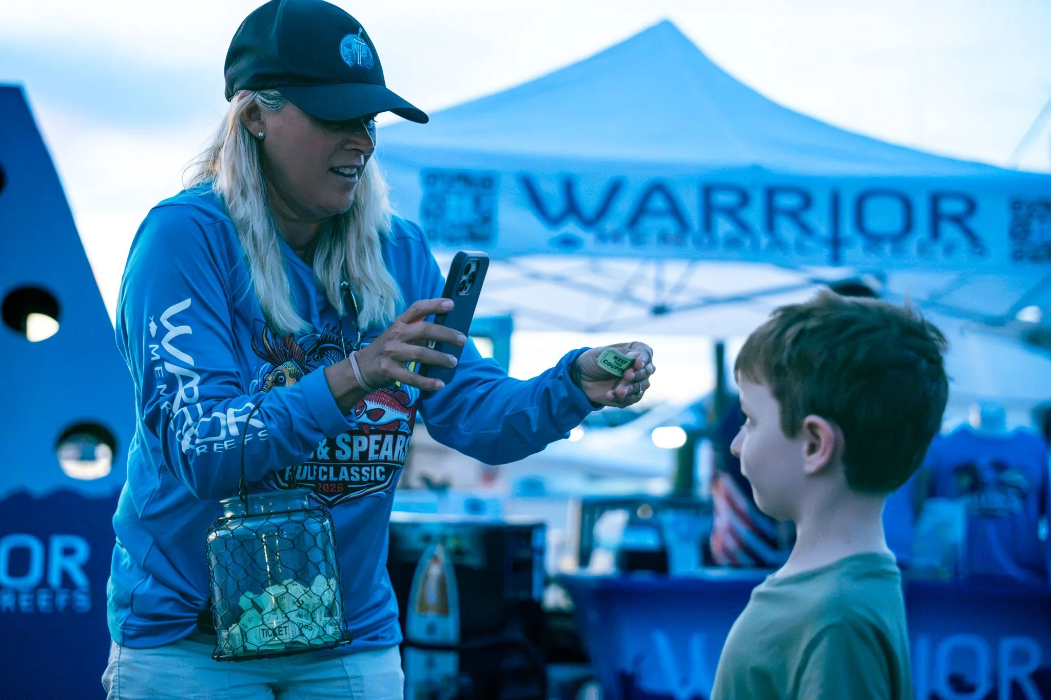 A woman wearing a black cap and blue sports jersey is holding a smartphone and engaging with a young boy with brown hair, outdoors at a sports or event tent.