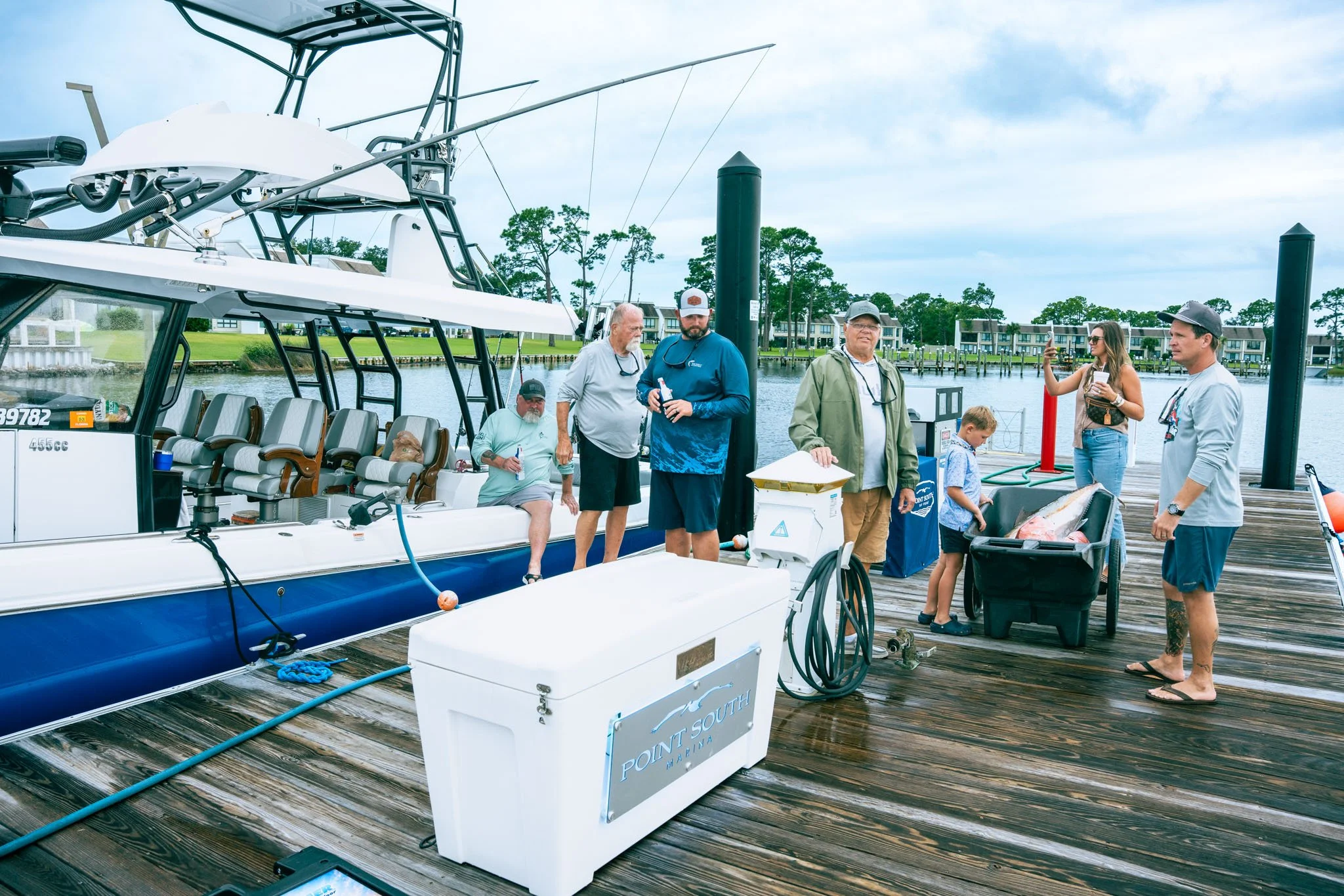 Group of people at Point South Marina - Bay Point (Panama City Beach) on a dock by a boat, with trees and water in the background, some holding drinks and taking photos, and a sign that reads 'Point South Marina'.