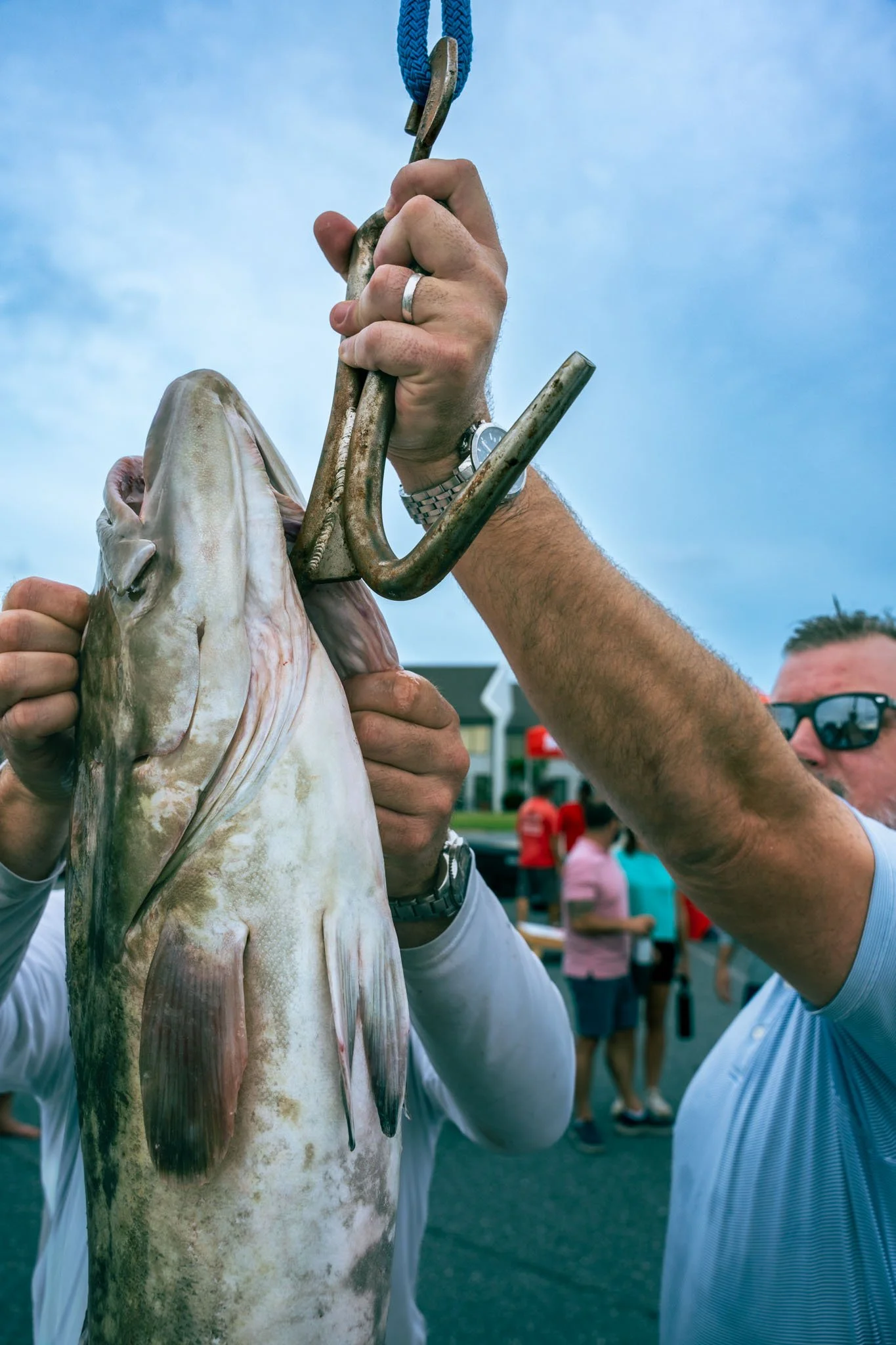 A person holding a large fish with both hands and a blue rope with a hook attached in the background. Other people are visible in the background, indicating an outdoor setting.