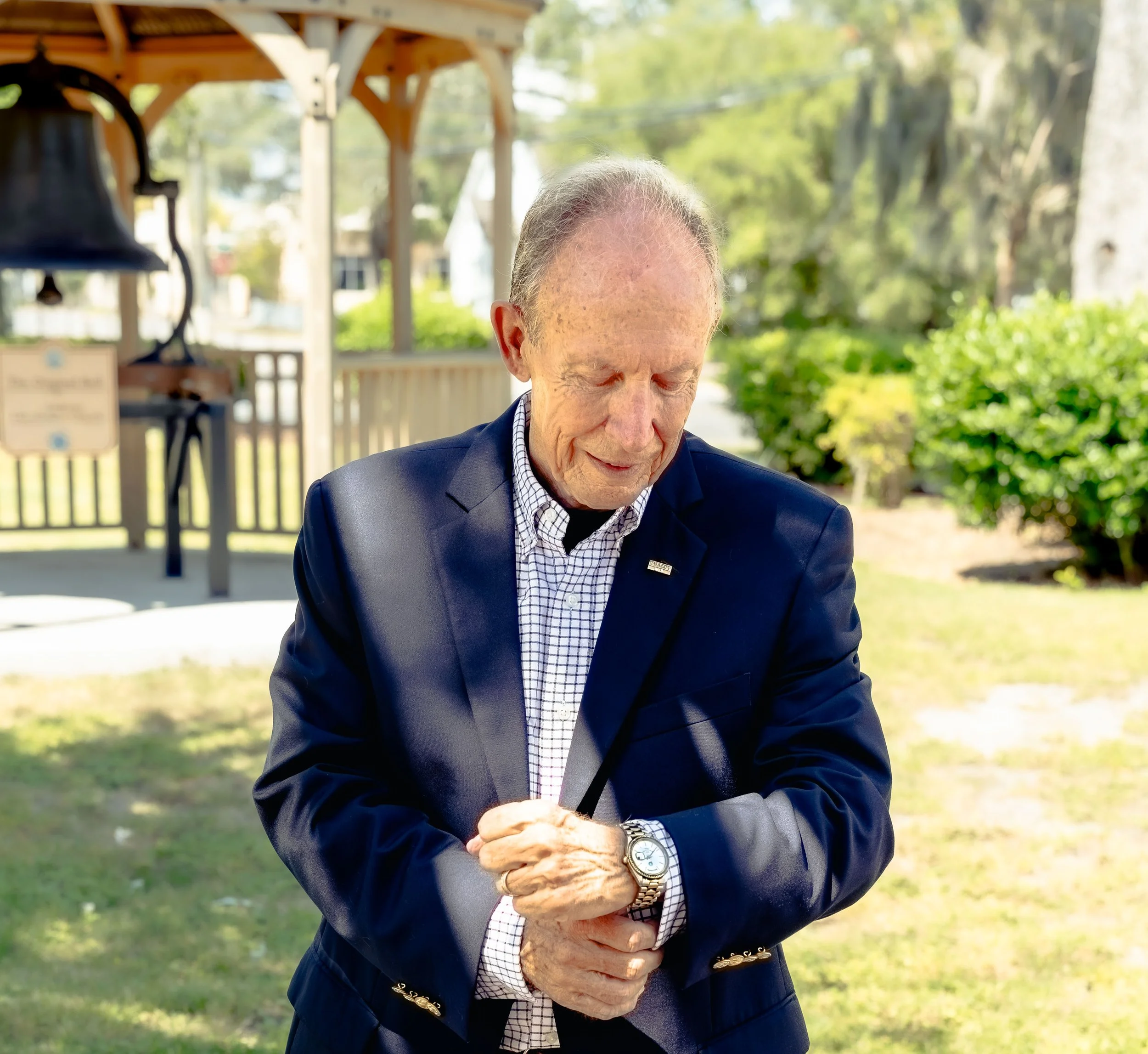 An elderly man with gray hair, wearing a navy blazer and a checkered shirt, looks down with his eyes closed outdoors in a sunny garden with green trees and a wooden structure in the background.