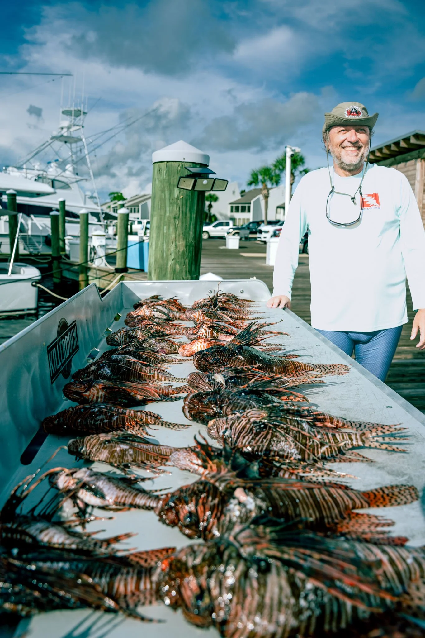 Man smiling at Point South Marina - Bay Point (Panama City Beach) with Lionfish on display on the cleaning station, boats and houses in the background under a blue sky with clouds.