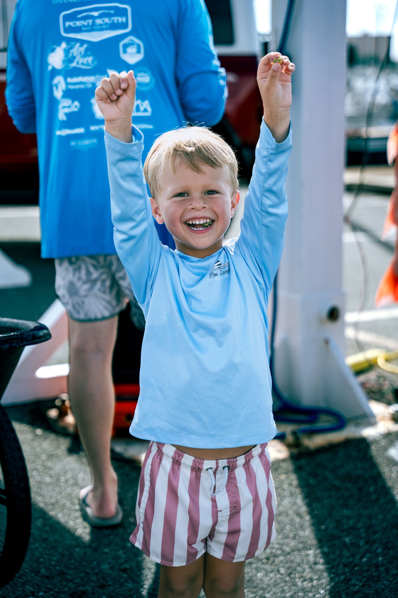 A young boy with blonde hair and a big smile, wearing a light blue long sleeve shirt and pink and white striped shorts, raising his arms in celebration outside.