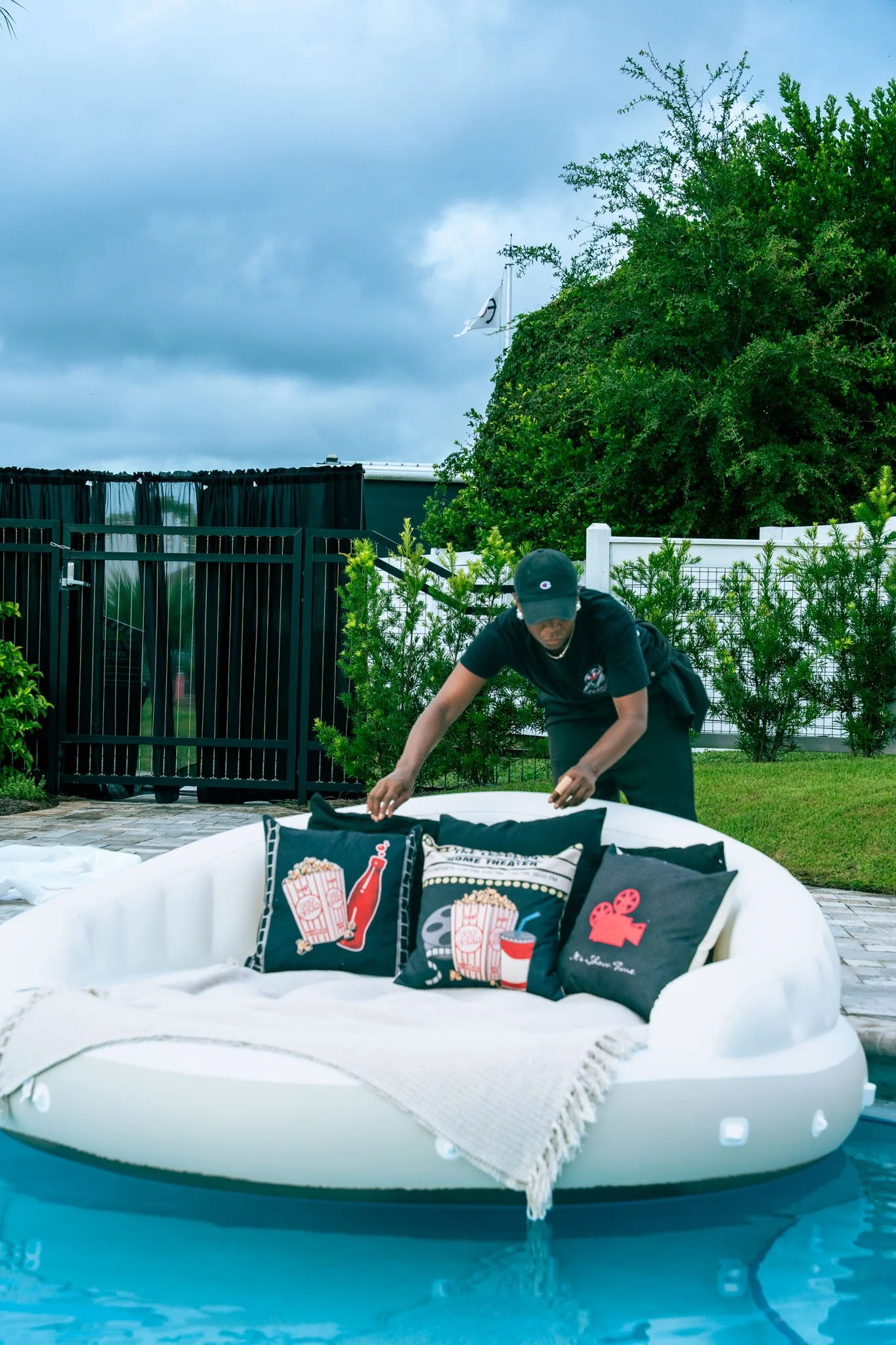 A person setting up decorations by a swimming pool, with a large inflatable pool float in the foreground and a table with themed pillows and snacks. There is a fenced yard with trees and a cloudy sky in the background.