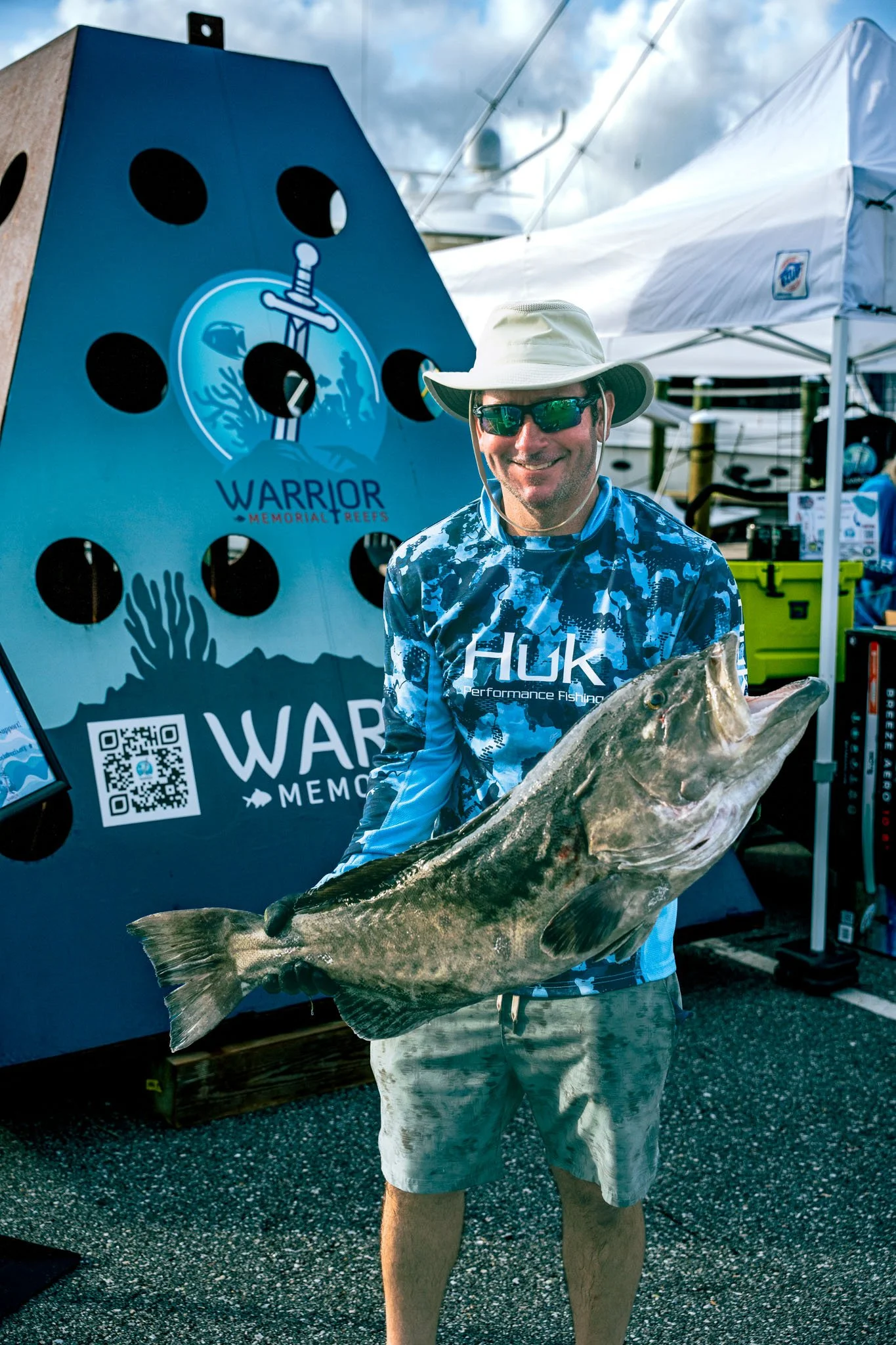 A man wearing a wide-brimmed hat, sunglasses, and a blue fishing shirt is holding a large fish, standing in front of a booth with a banner that reads 'Warrior Memorial Reefs' and displays a sword and coral illustration, at an outdoor event near boats