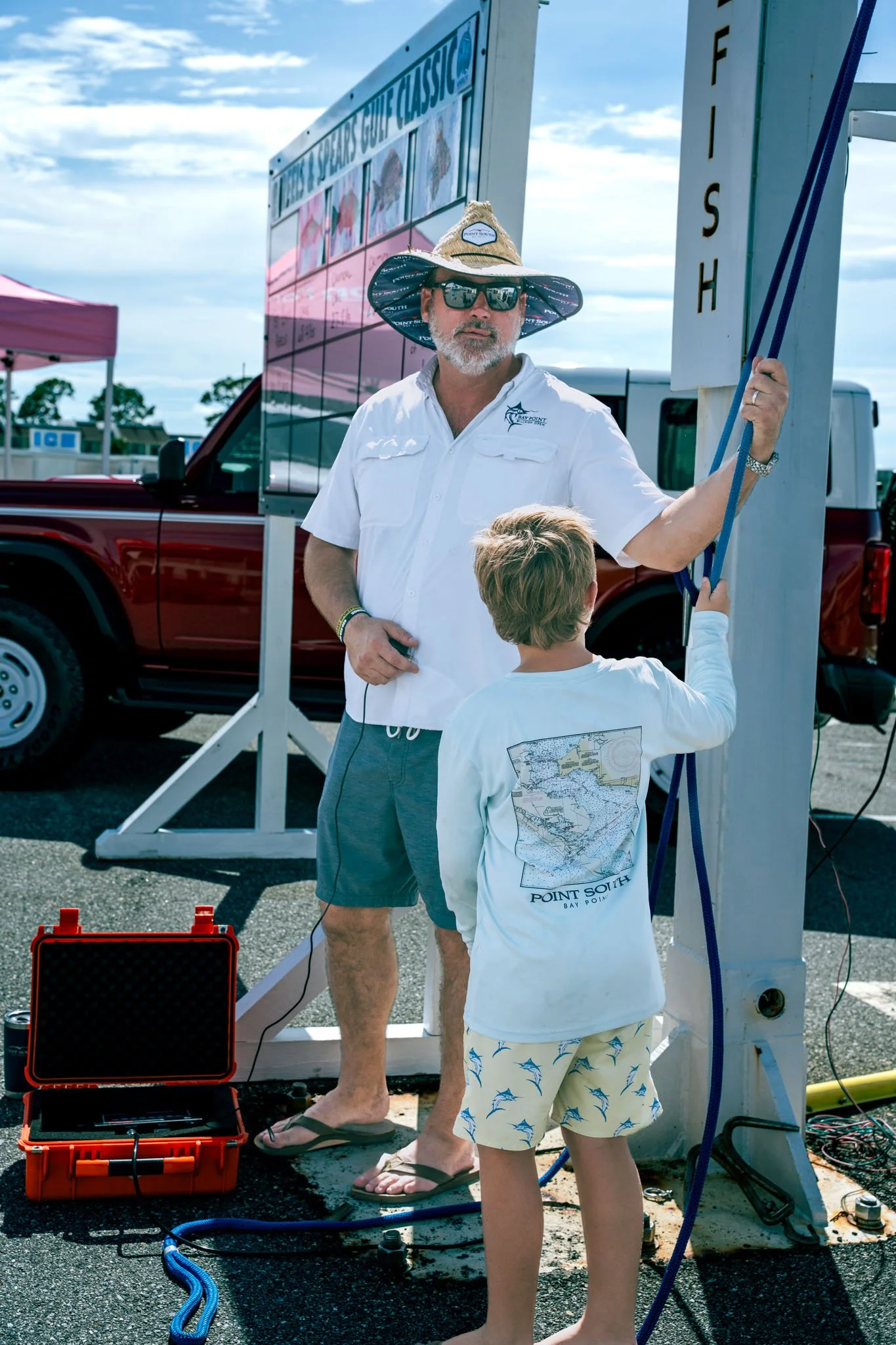A man with a beard, wearing sunglasses, a wide-brimmed hat, a white shirt, and shorts, standing next to a young boy dressed in shorts and a long-sleeve shirt, both facing and working on a white wooden structure with blue ropes. There are racks and tr