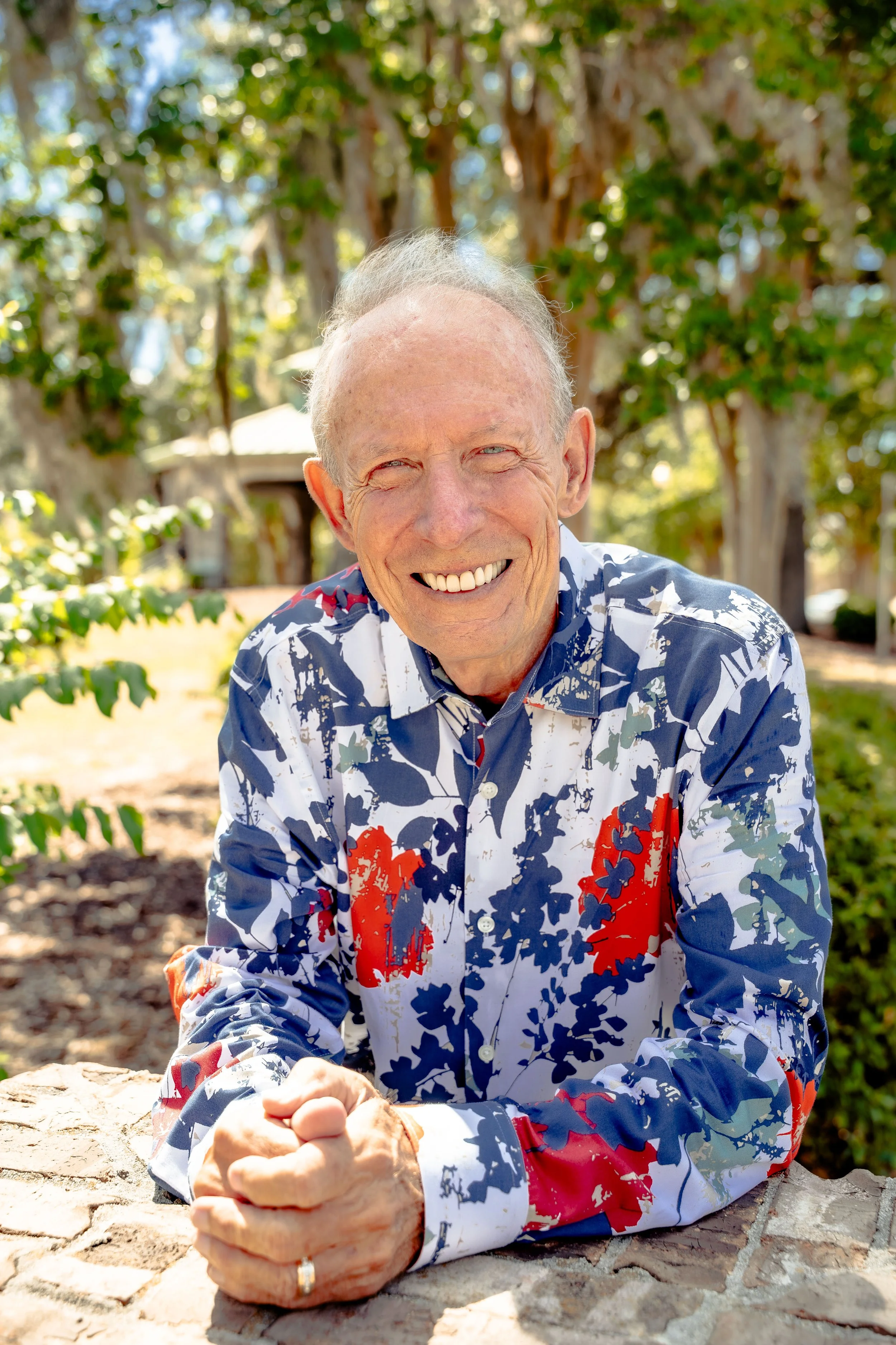 A smiling elderly man with gray hair and blue eyes, wearing a colorful Hawaiian shirt, sitting outdoors during daytime, with trees in the background.