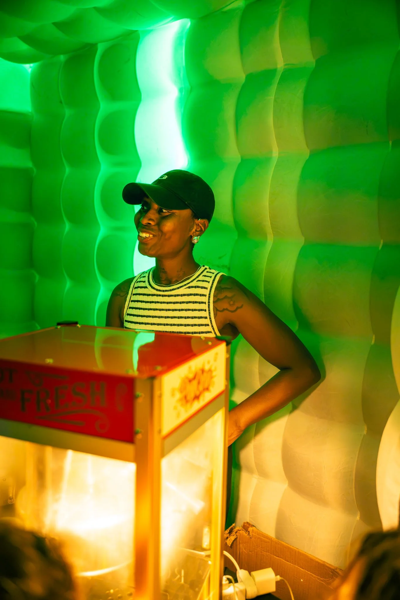A woman with a dark cap, earrings, and a sleeveless striped top, smiling, stands against a textured green wall.