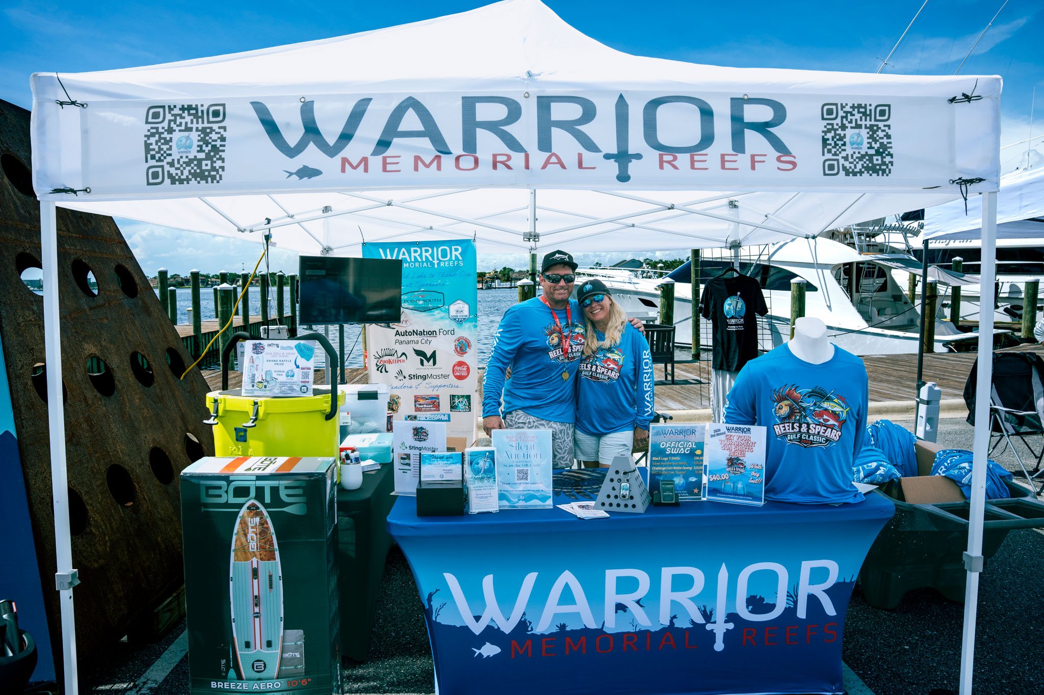 A promotional booth for Warrior Memorial Reefs at a marina, featuring a man and woman wearing blue T-shirts with a fishing graphic, standing behind a table with merchandise, signs, and a tent with the Warrior Memorial Reefs logo and QR codes, boats a