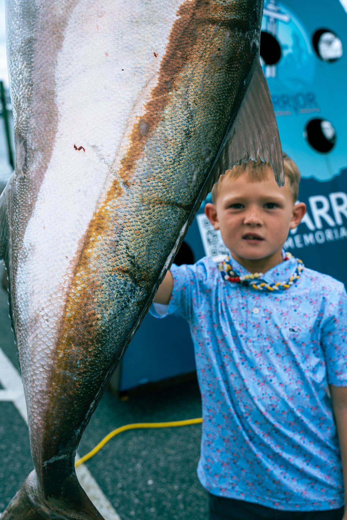 A young boy with short blonde hair wearing a blue floral shirt and beaded necklace, standing outdoors next to a large fish with shiny, scaled skin. The boy is holding the fish up, partially obscured by the fish.