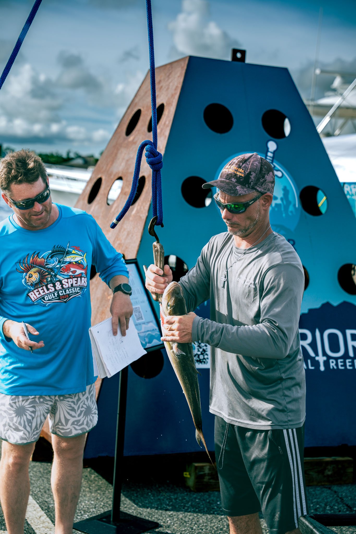 Two men are holding a fish in front of a large blue and wooden structure with black circular holes. One man is wearing sunglasses, a blue shirt with colorful graphics, and patterned shorts, while the other man is wearing sunglasses, a cap, a gray shi