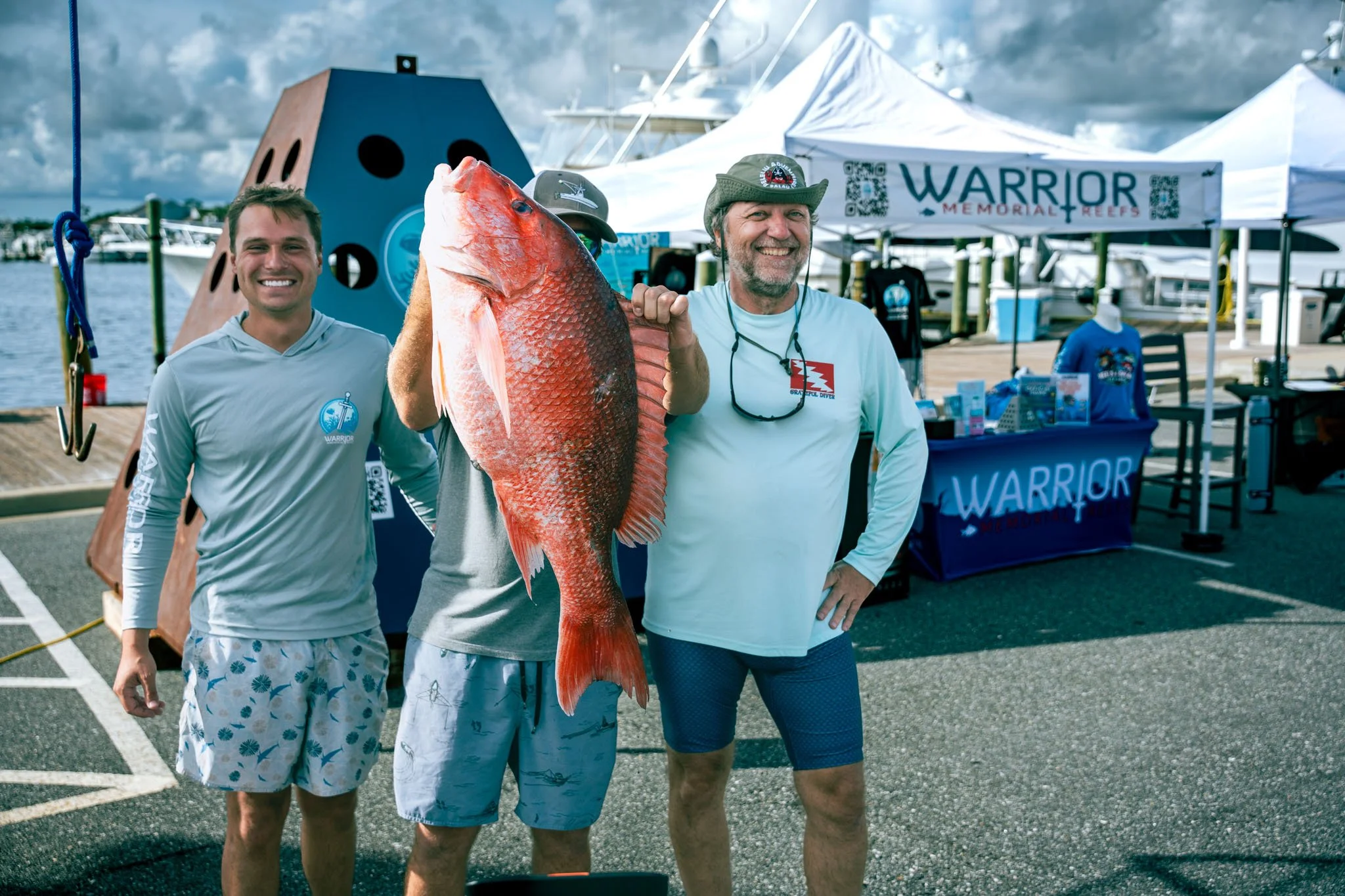 Three men standing in a parking lot near boats and tents, with one holding a large red fish. They are smiling and appear to be at a fishing event or festival, with a banner in the background reading 'WARRIOR Memorial Reefs'.