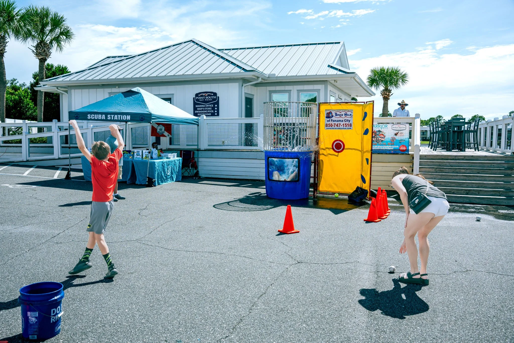 Children playing shuffleboard outdoors near a white building with a metal roof. There is a table under a blue tent with the words 'Scuba Station' on it, and a yellow barrier with a Target logo. A person stands on the deck of the building while palm t