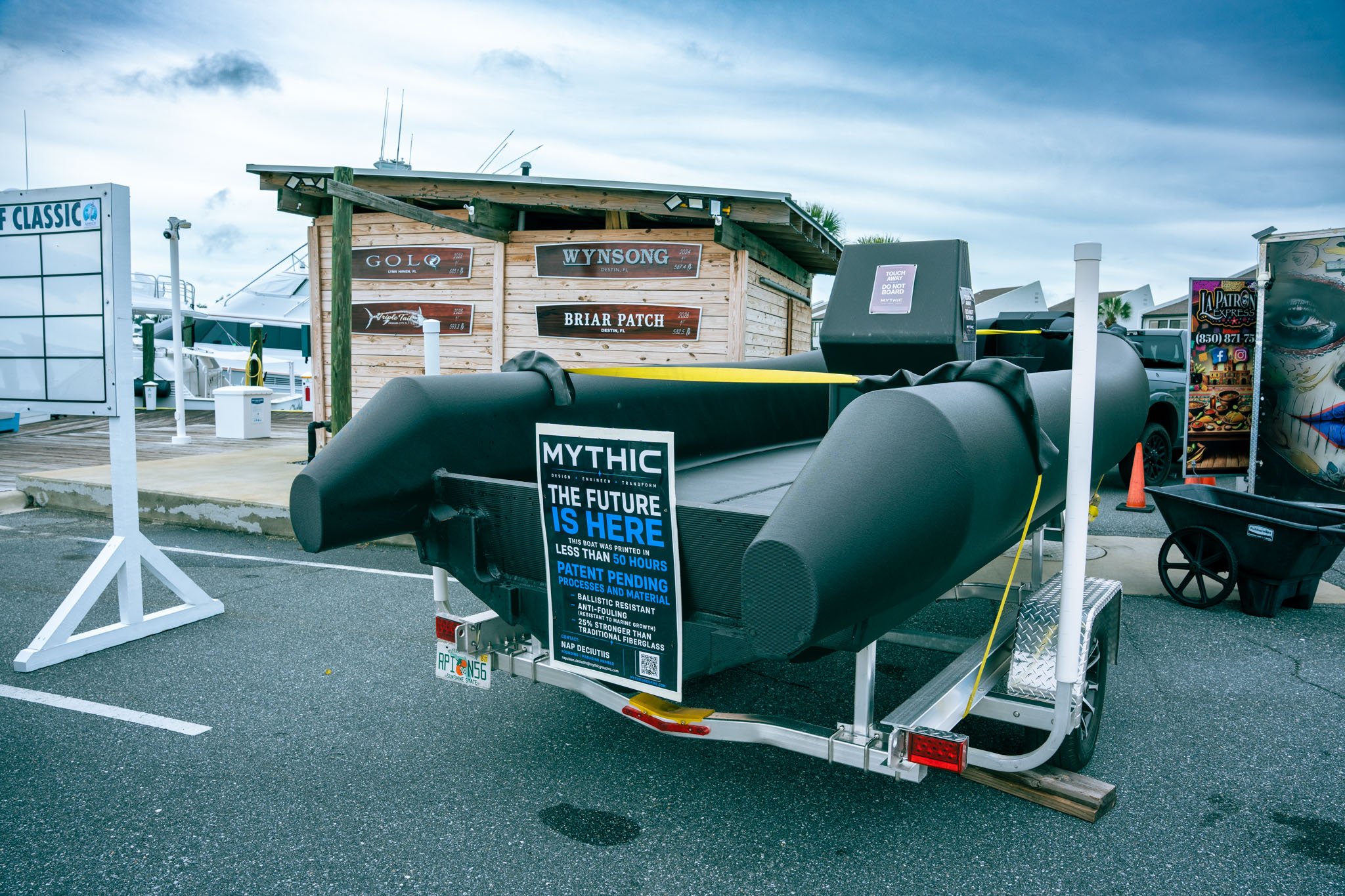 View of a boat trailer with a sign attached, docked at Point South Marina - Bay Point (Panama City Beach) with boats and a wooden shed in the background, under a cloudy sky.