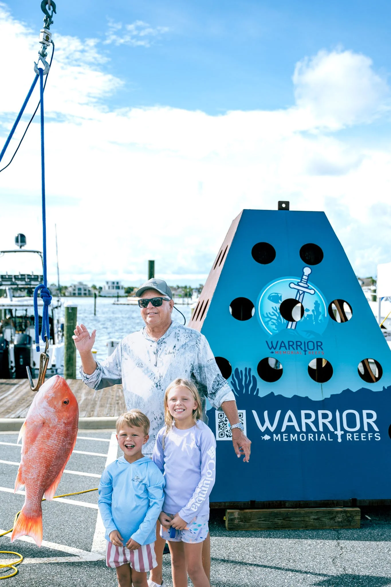 A man and three children standing on a dock with a large fish and a blue Shark Memorial Reef sign behind them at Point South Marina - Bay Point (Panama City Beach). The man is waving, wearing a cap and sunglasses, and the children are smiling.
