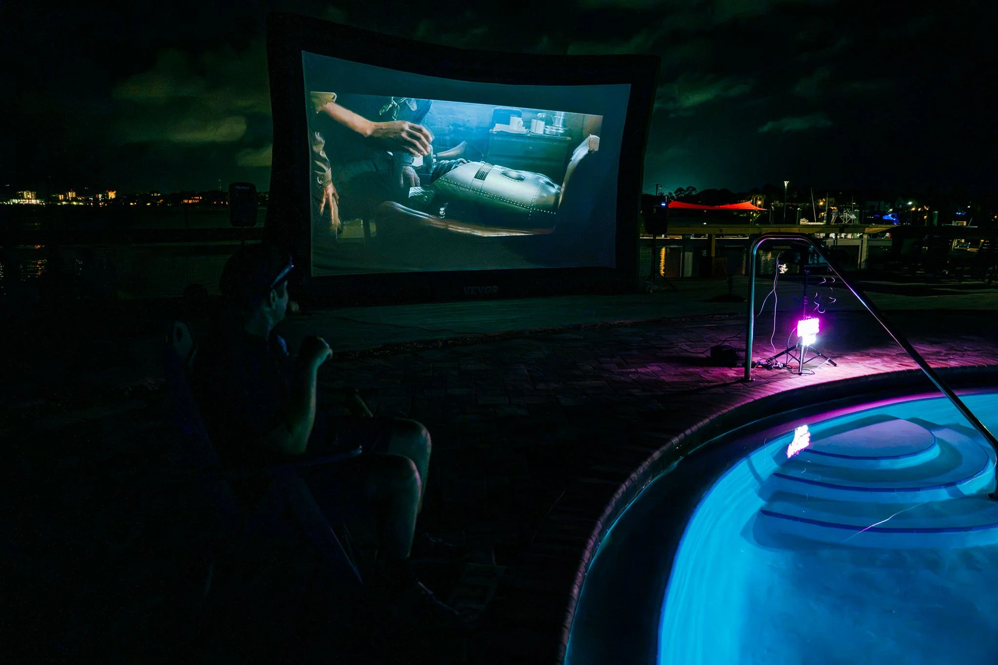 Night scene of an outdoor movie screening with a large projection screen showing a scene of a person in a cockpit, with people sitting nearby watching the film alongside Point South Marina - Bay Point (Panama City Beach) pool with illuminated water.