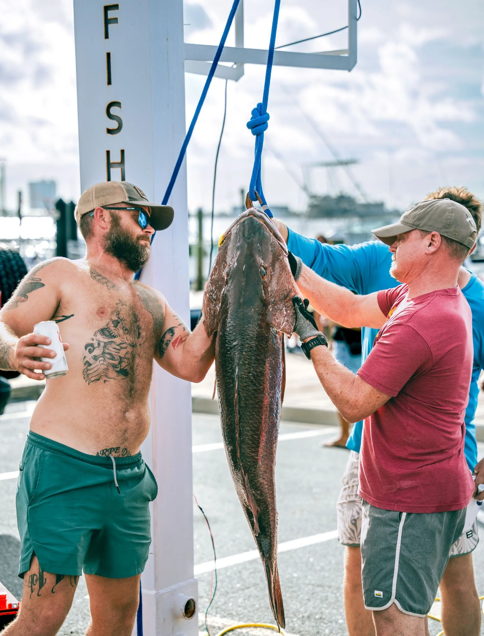 Two men are preparing a large fish, likely a shark, for display or measurement outdoors near a marina, with boats in the background. One man is shirtless with tattoos and wearing sunglasses and shorts, holding a beverage, while the other man is weari