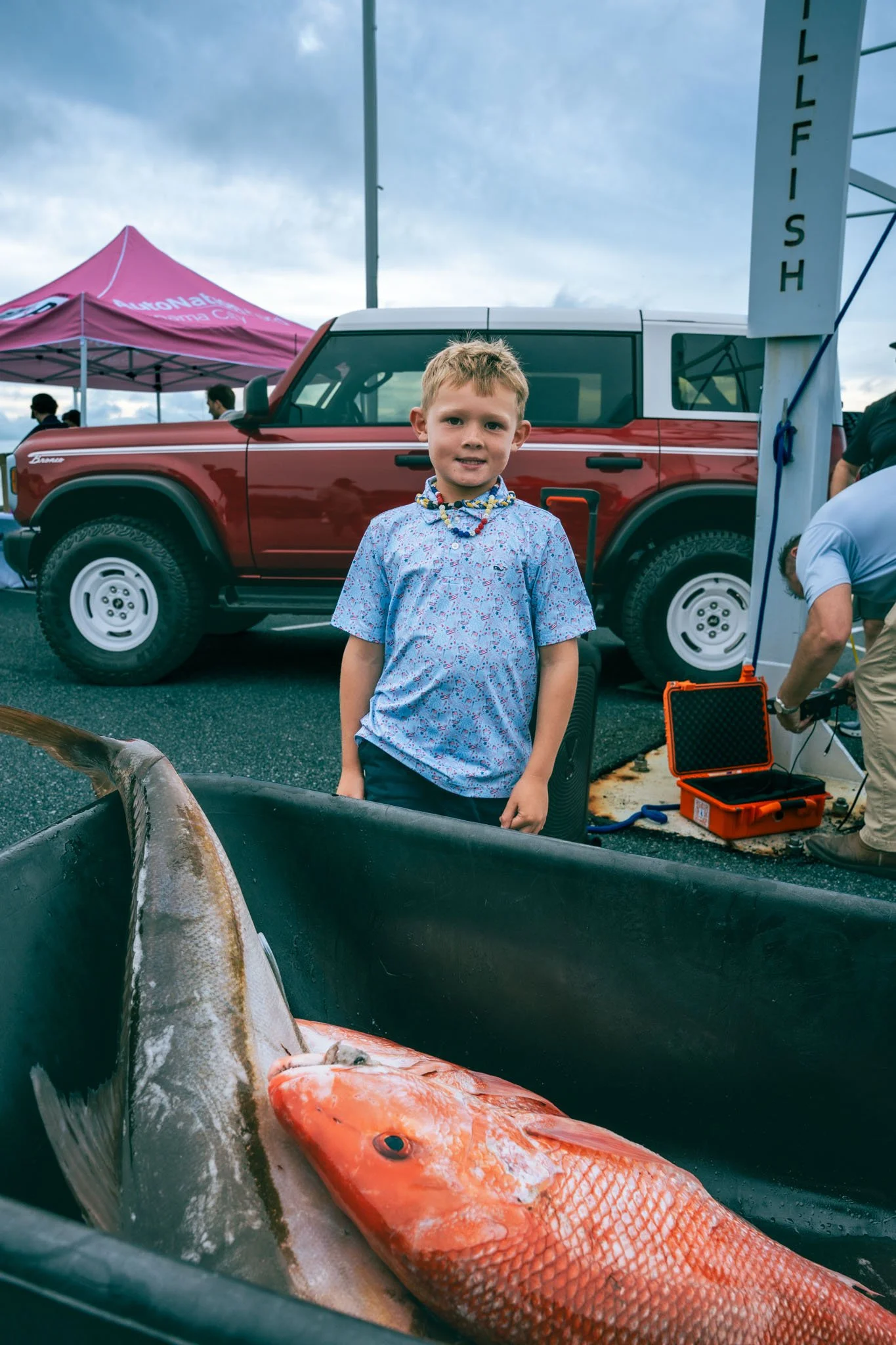 A young boy standing in front of a red truck at an outdoor fishing event at Point South Marina - Bay Point (Panama City Beach), with large fish lying in a black container in the foreground.