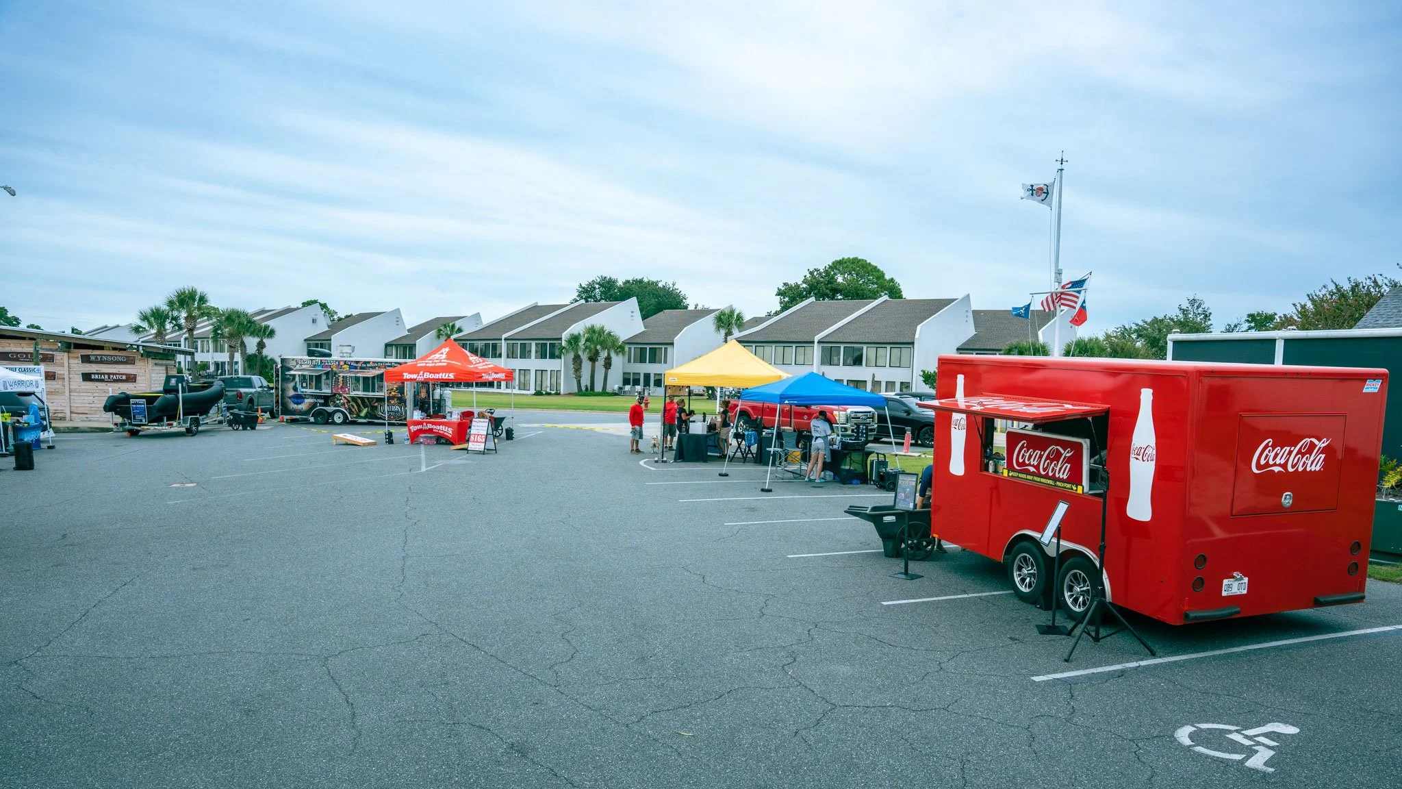 Outdoor parking lot at Point South Marina - Bay Point (Panama City Beach) with food and drink vendors, including a Coca-Cola truck and traditionale tents, with modern buildings and trees in the background under a cloudy sky.