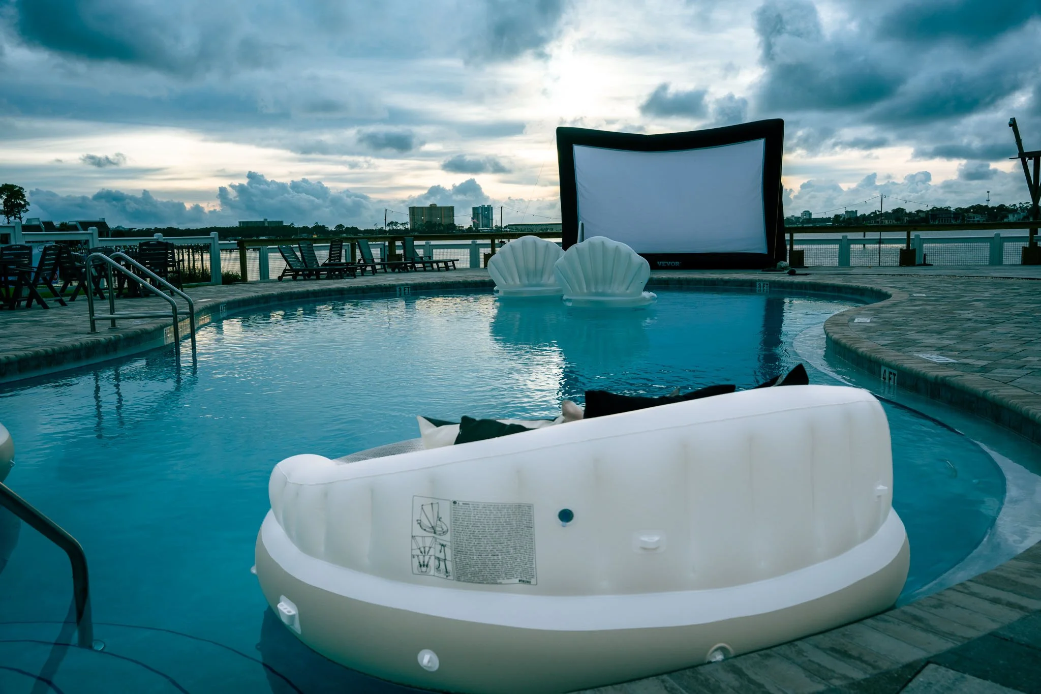Indoor pool area with pool float, lounge chairs, and a large screen or projector, with a city skyline and cloudy sky in the background.