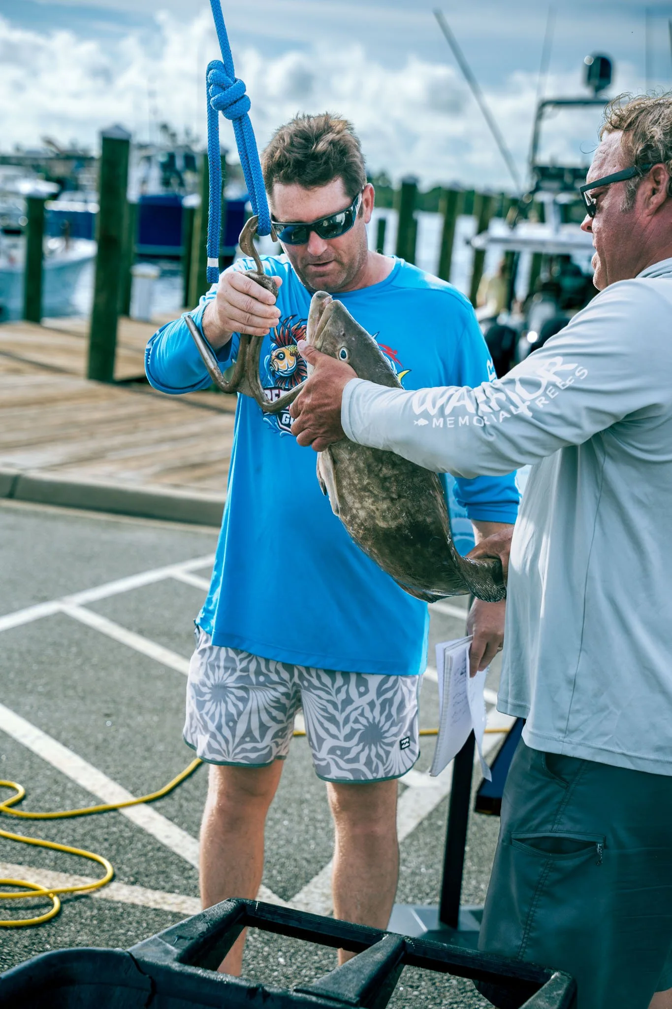 Two men holding a fish on a dock at Point South Marina - Bay Point (Panama City Beach), with boats and cloudy sky in the background.